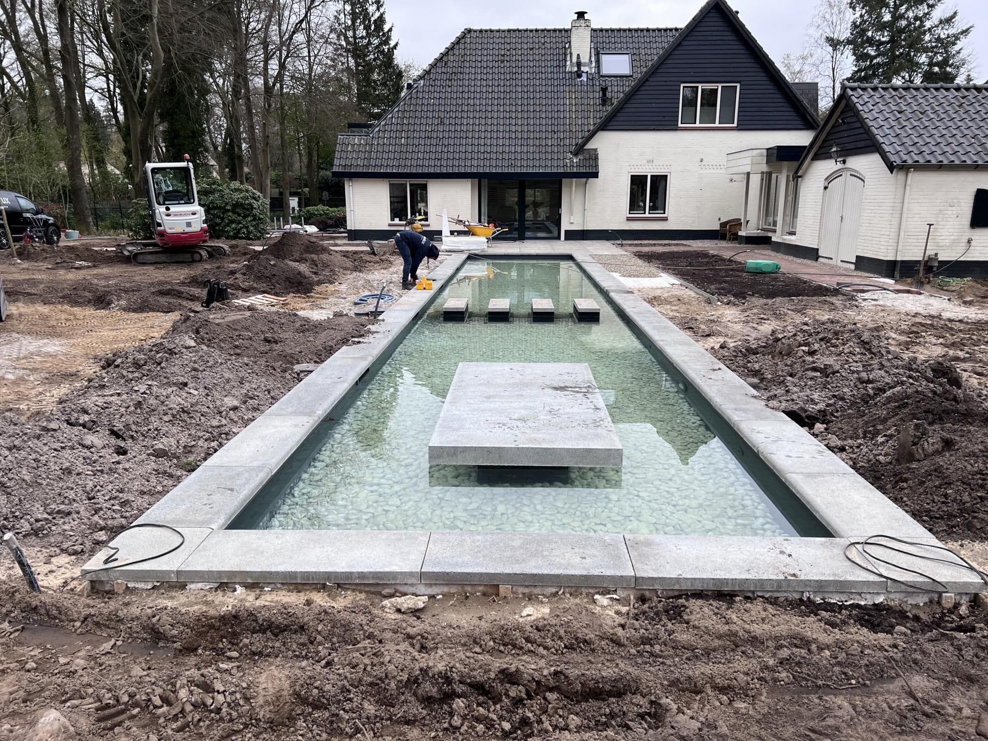 Rechthoekig zwembad met water en betonnen stapstenen in aanleg; rondom graafwerk, grondhopen, een graafmachine en een witte woning.