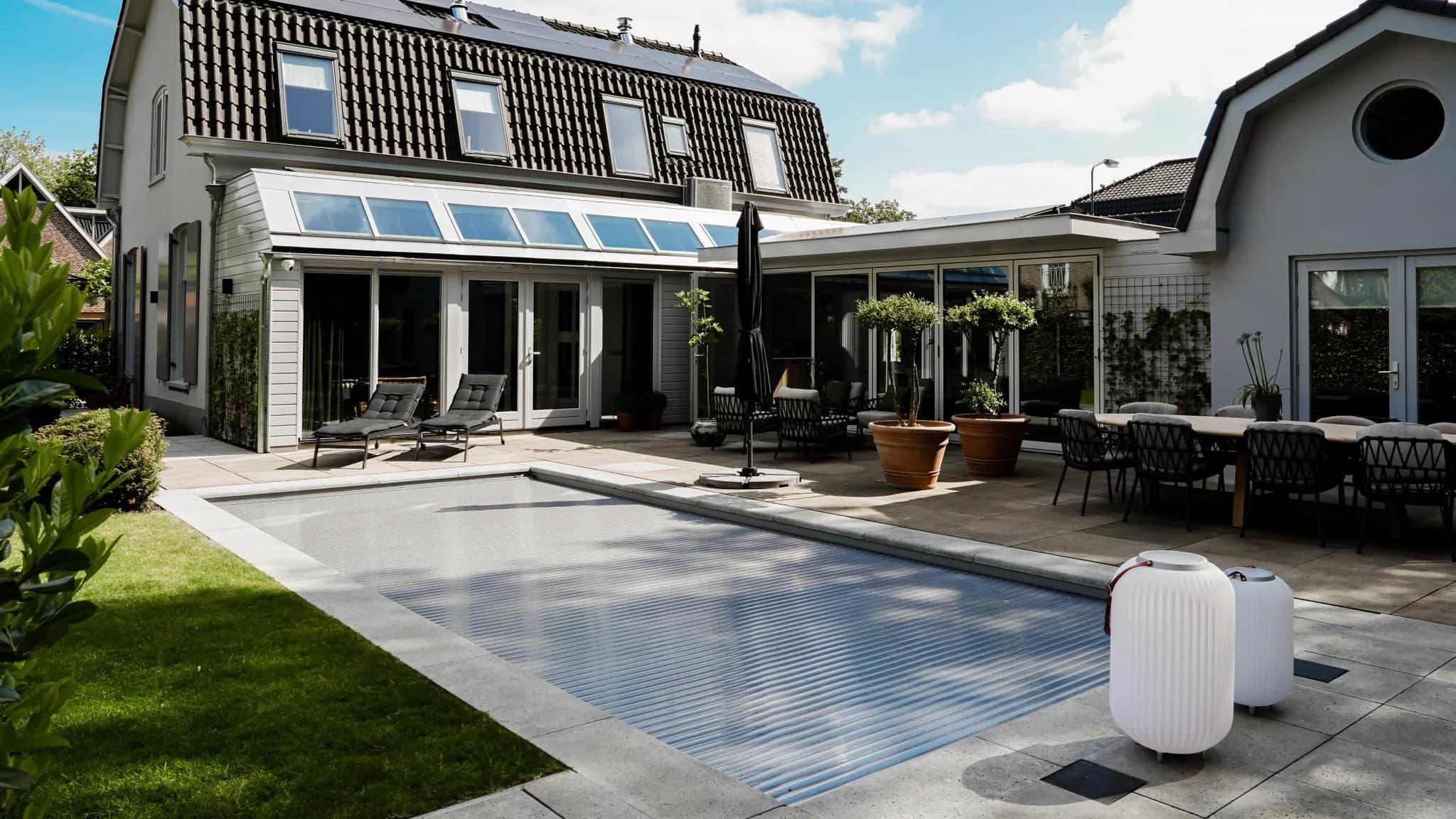 Terras en achtertuin van een modern huis met ligstoelen, planten en een zwembad met rolluikafdekking voor grote glazen schuifdeuren.