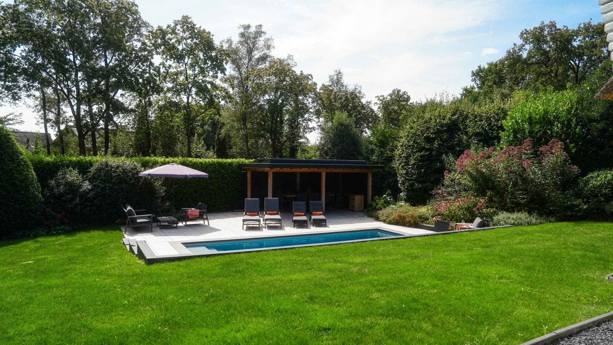 Achtertuin met groen gazon, rechthoekig zwembad op een betegeld terras, vier ligstoelen met oranje accenten, parasol en een houten overkapping, omringd door hagen en hoge bomen.