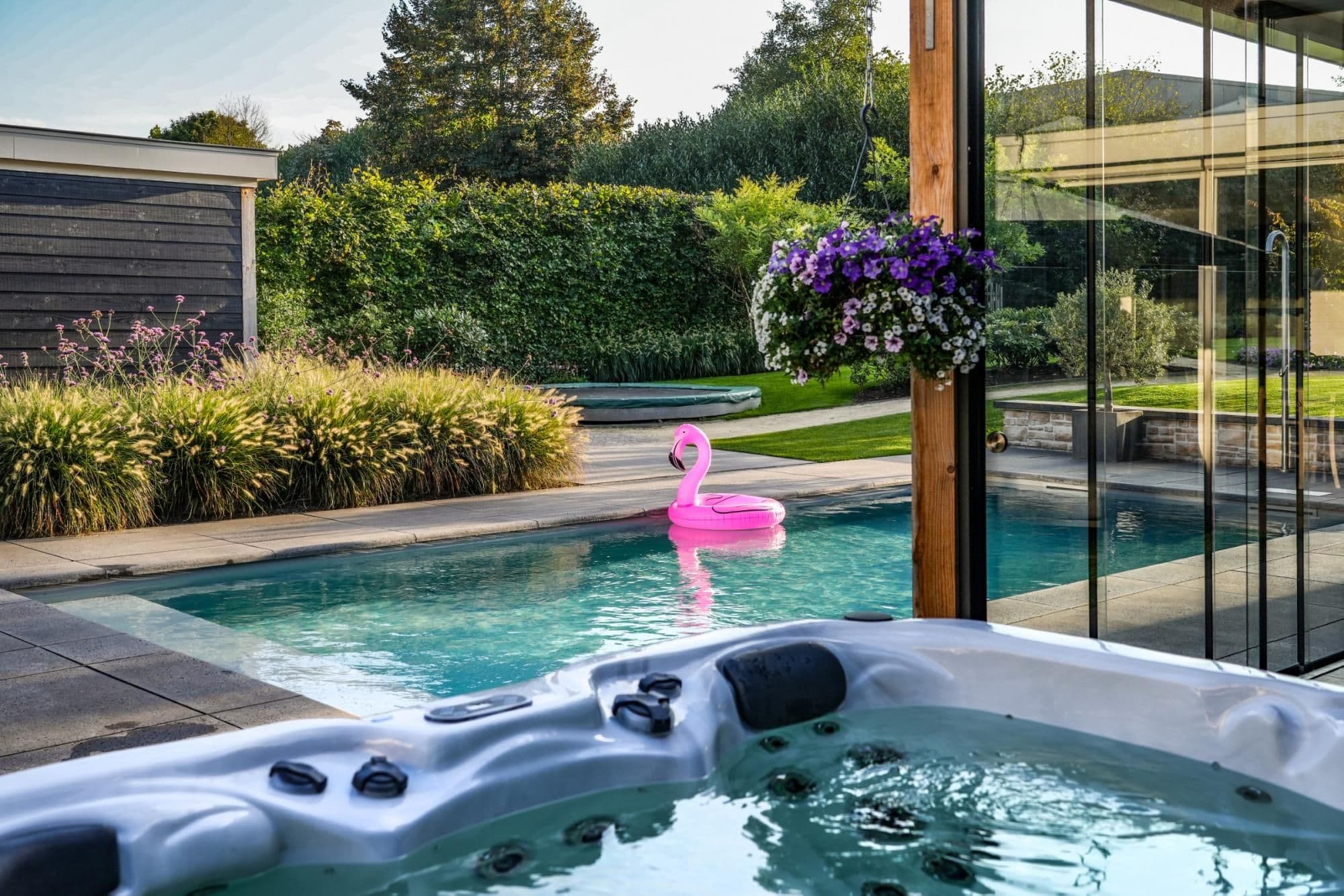 Zwembad in achtertuin met roze opblaasflamingo, op de voorgrond een bubbelbad en rechts glazen schuifdeuren en een hangende bloembak met paarse bloemen.