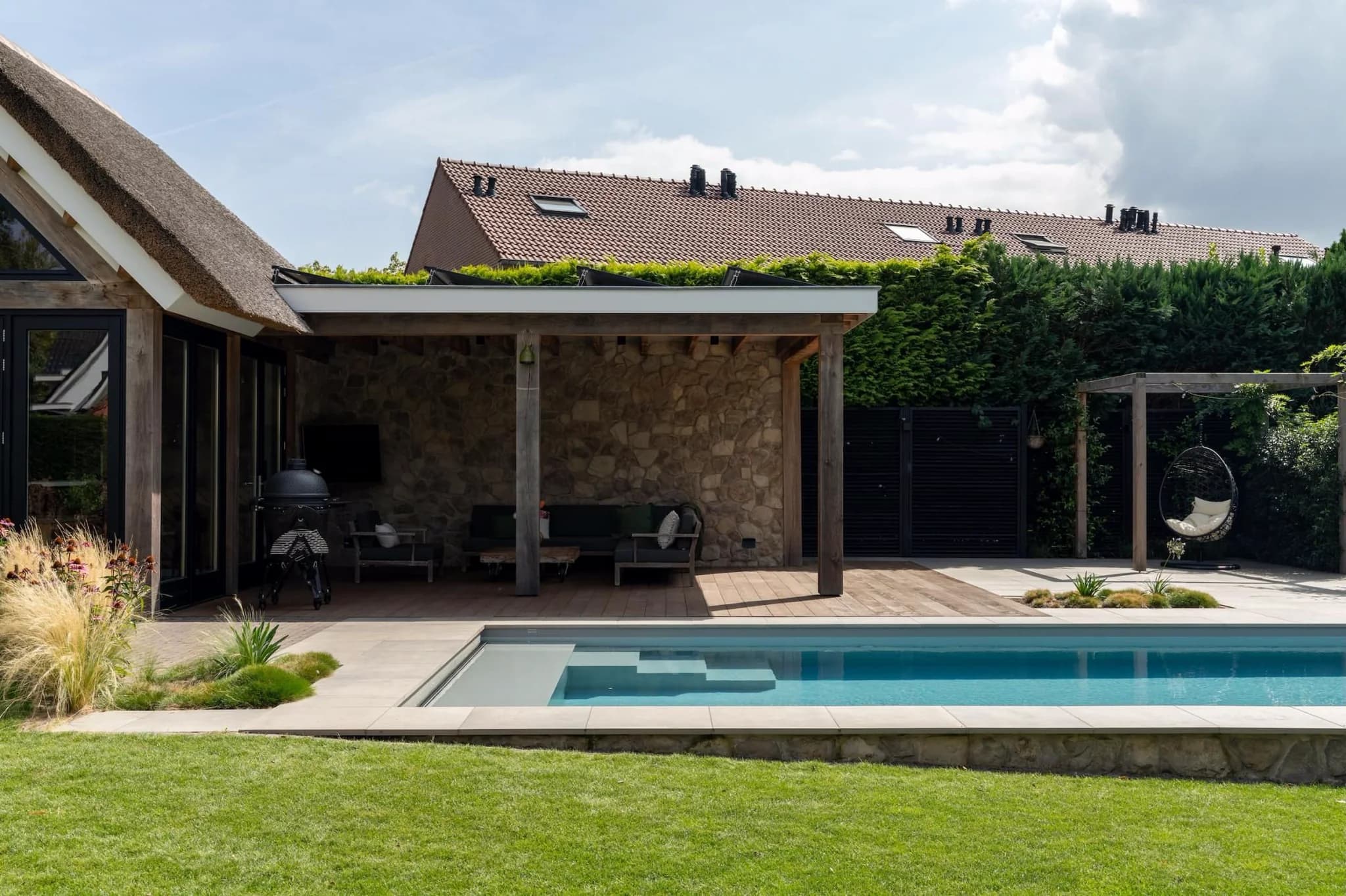 Zwembad met trap in voorgrond, overdekt terras met houten palen en stenen muur in het midden, links rieten dak en glazen deuren, rechts hoge haag en hangstoel