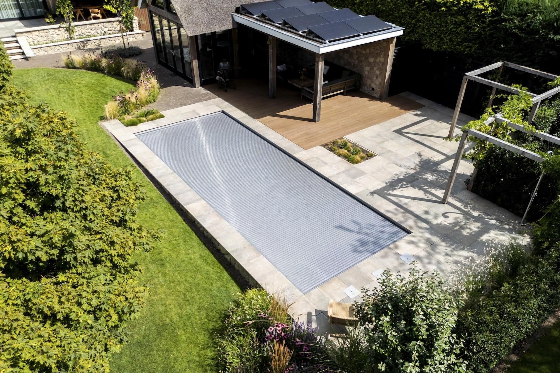 Luchtfoto van achtertuin met rechthoekig zwembad met gesloten rolafdekking, betegeld terras, houten overkapping met zonnepanelen en omliggende planten
