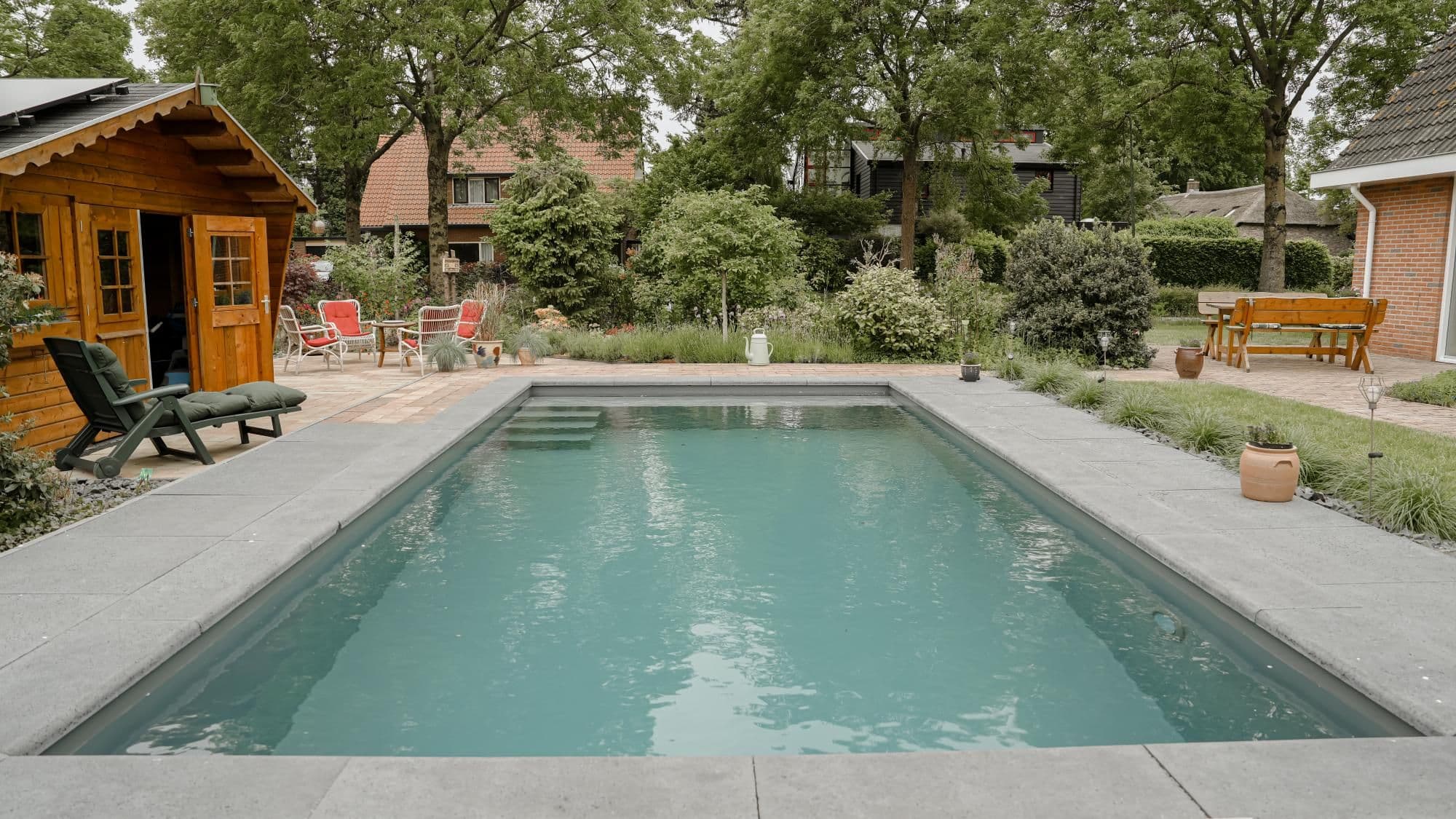 Rechthoekig zwembad in een achtertuin met een houten tuinhuis links, ligstoel, tuinmeubels en veel beplanting