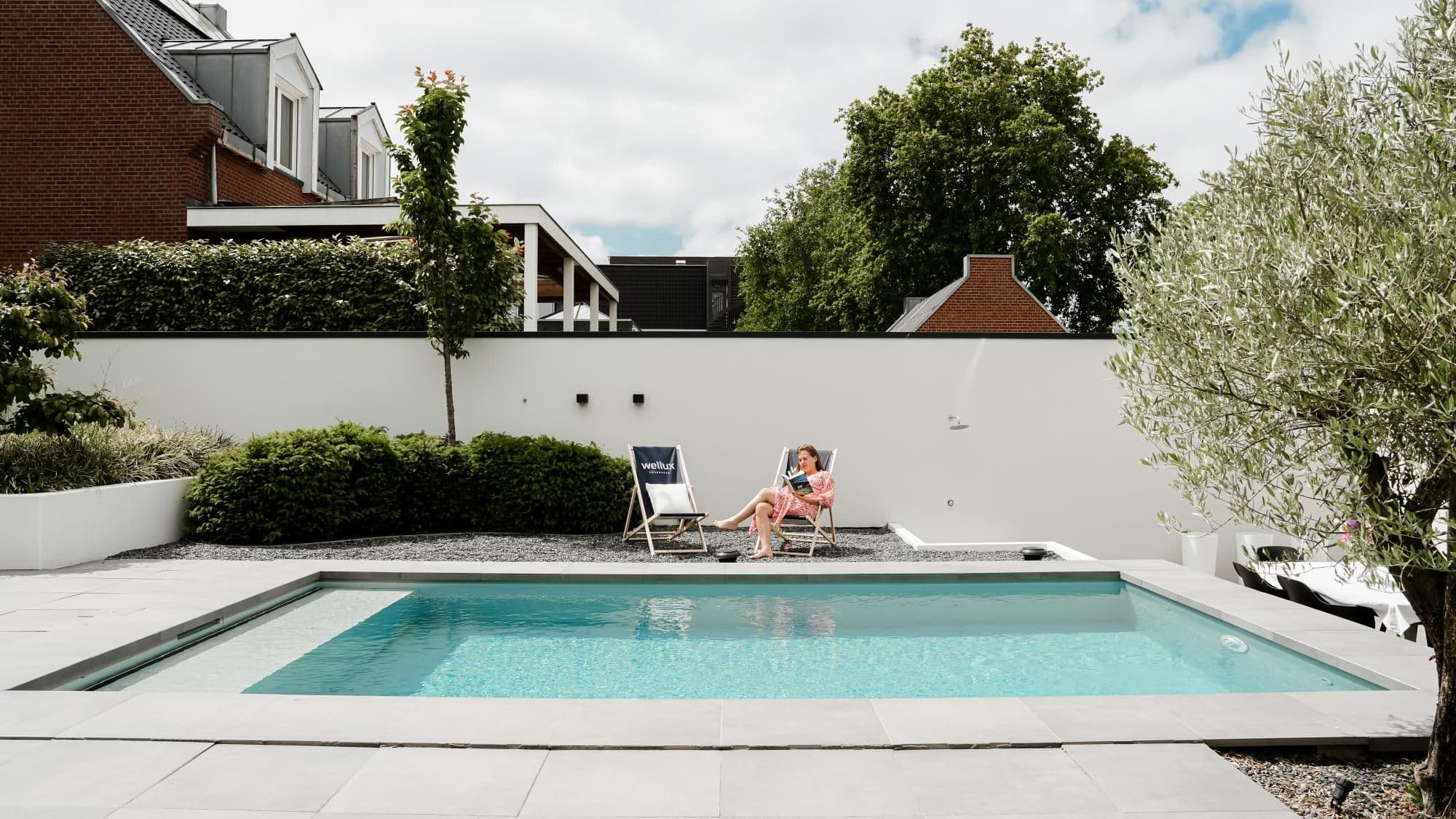 Rechthoekig zwembad op de voorgrond, witte tuinmuur met twee ligstoelen rechts van het midden, vrouw leest op een ligstoel; struiken en bomen en bakstenen huizen op de achtergrond.