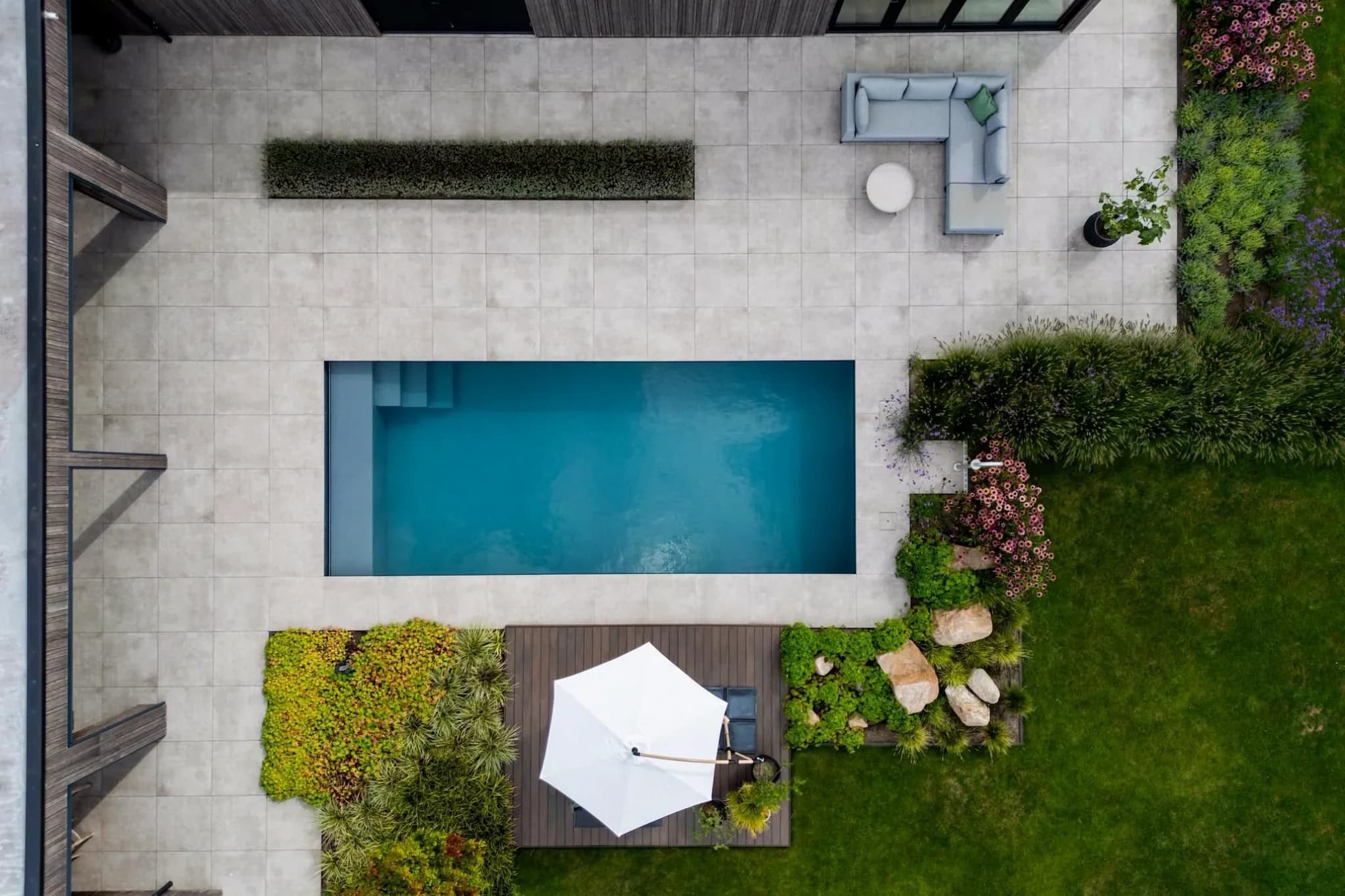 Luchtfoto van rechthoekig zwembad op betegeld terras met L-vormige loungeset, ronde tafel, houten vlonder met witte parasol en groene borders