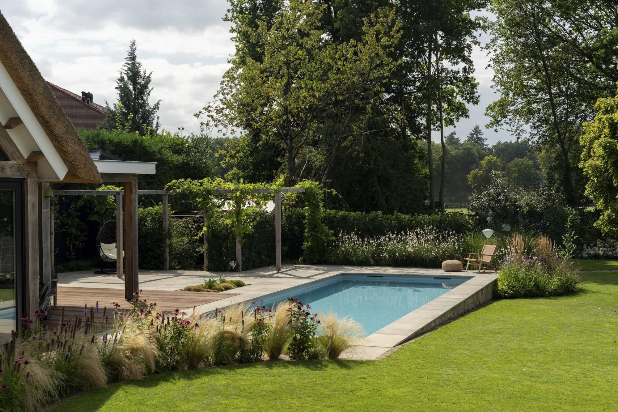 Rechthoekig buitenzwembad naast een houten terras met pergola, beplanting en een ligstoel, omgeven door gras en bomen