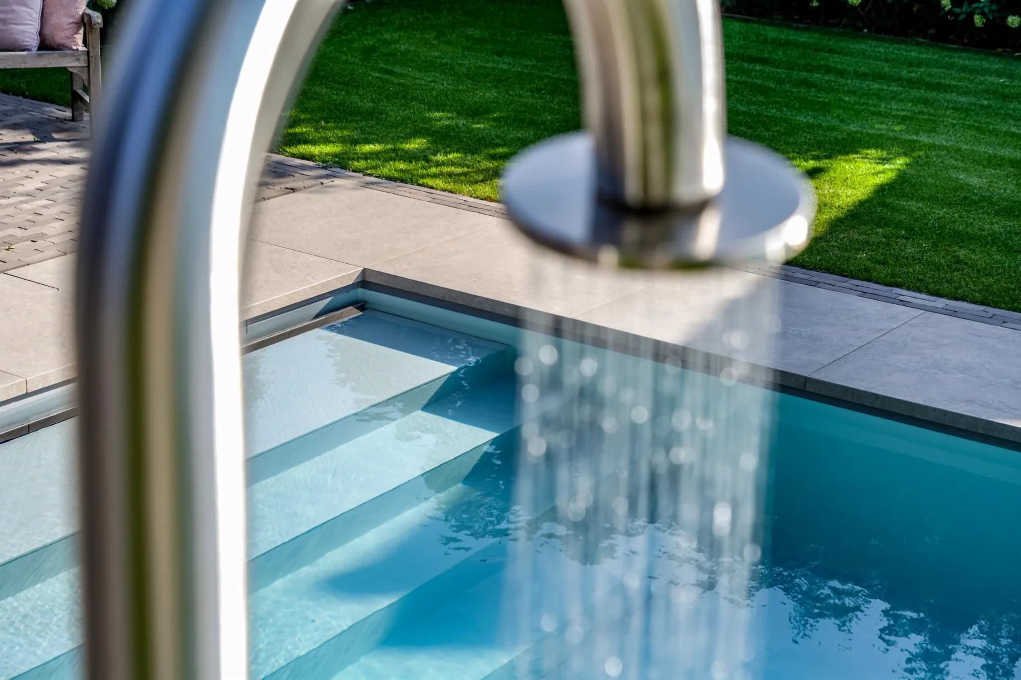 Close-up van een buitenzwembad met inlooptrap, stromend water uit een metalen douchekop en betegeld terras met gazon.