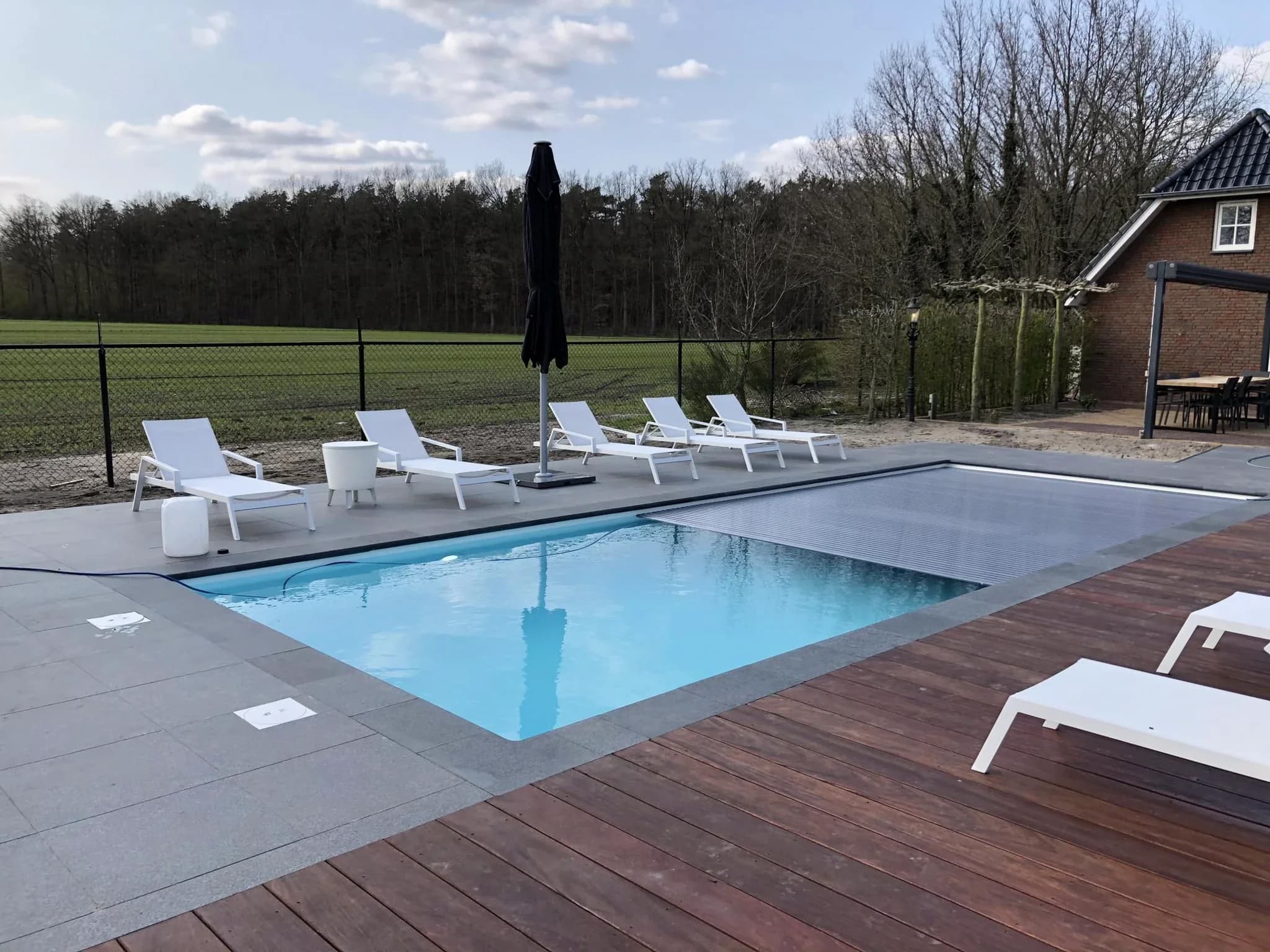 Zijaanzicht van een buitenzwembad met lichtblauw water en gedeeltelijke afdekking, rijen witte ligstoelen, een opgevouwen zwarte parasol op een betegeld terras naast een houten vlonder en een huis, met bomen en een veld op de achtergrond.