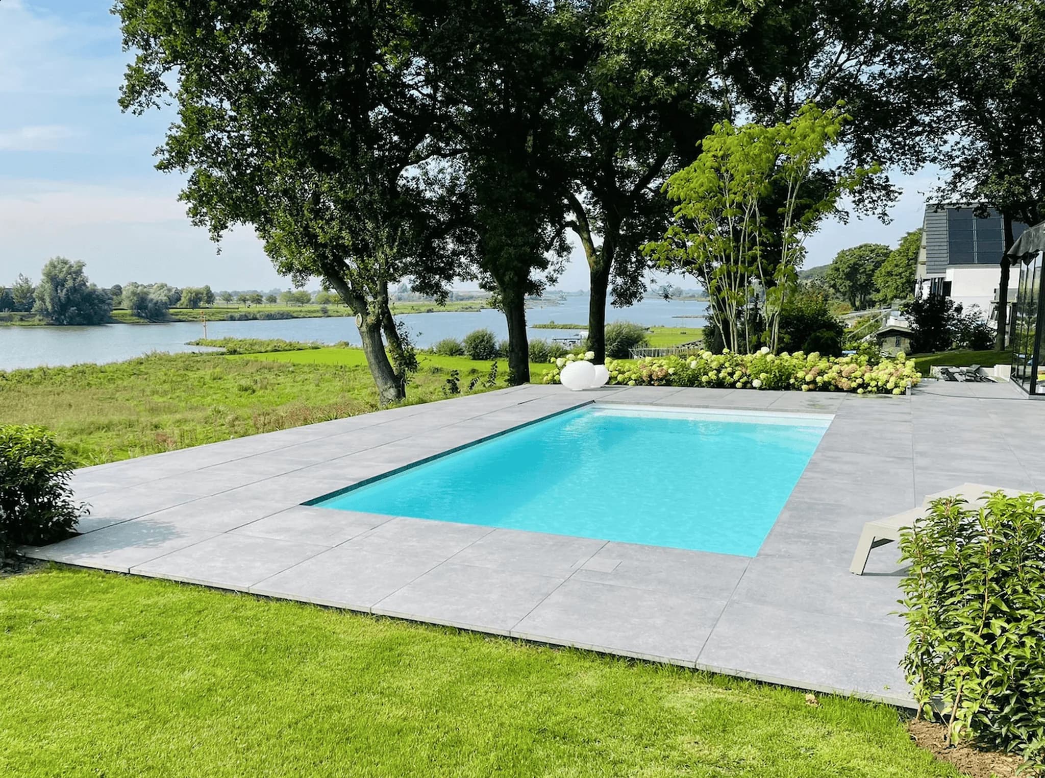 Rechthoekig buitenzwembad met helderblauw water en grijze terrastegels, omgeven door groen gazon, bloeiende struiken en hoge bomen; op de achtergrond een brede rivier en rechts een woning.
