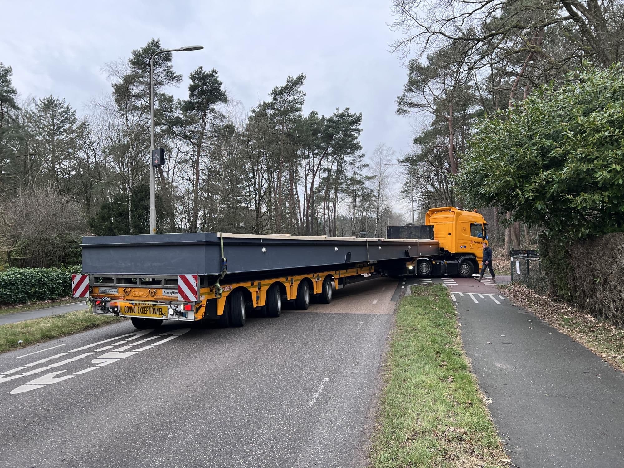 Gele vrachtwagen met lange oplegger op bochtige weg naast fietspad en bomen