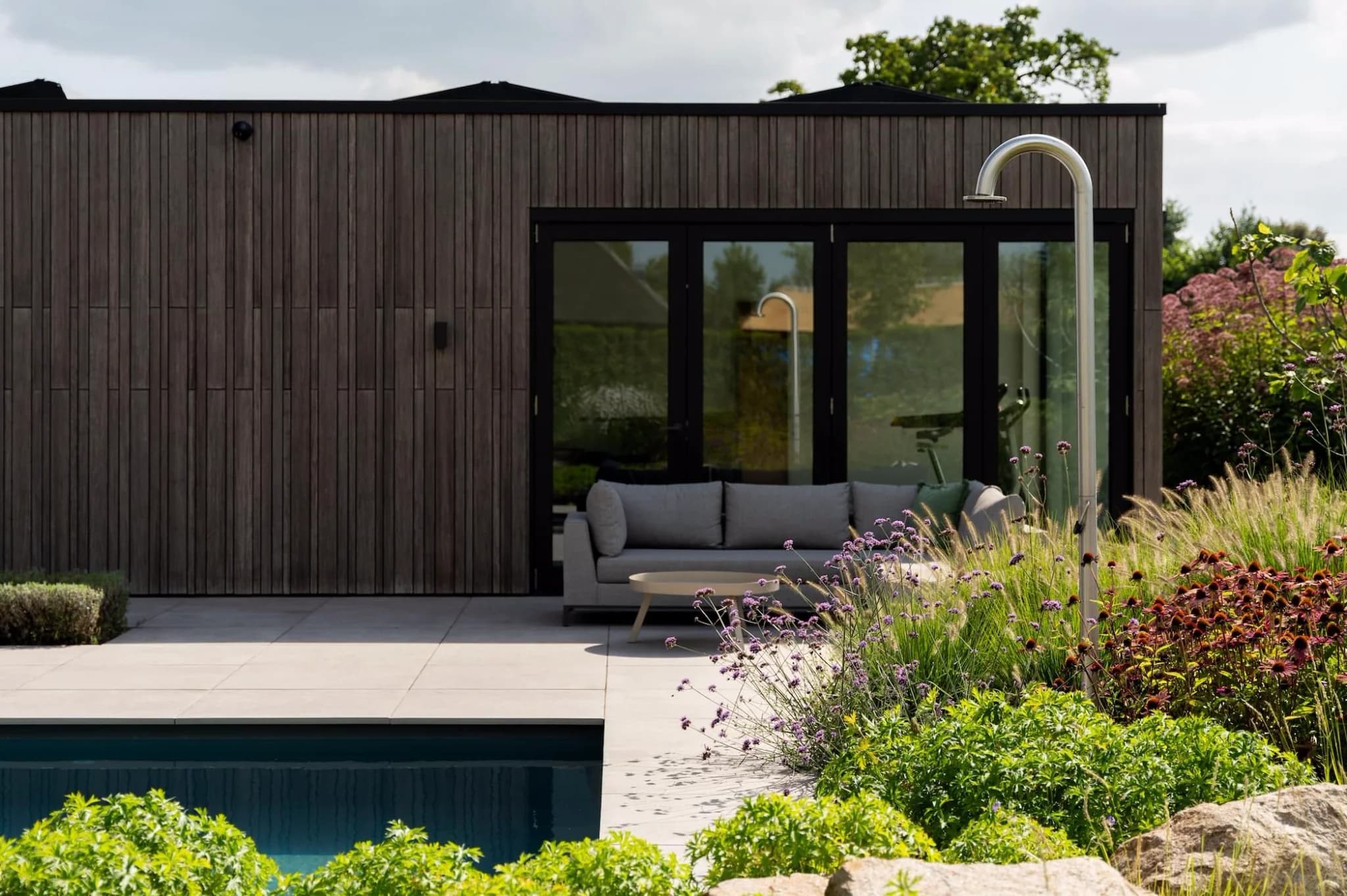 Terras met verticale houten gevel en zwarte glasdeuren, grijze sofa met lage tafel, roestvrijstalen buitendouche rechts en zwembad met voorgrondbeplanting.