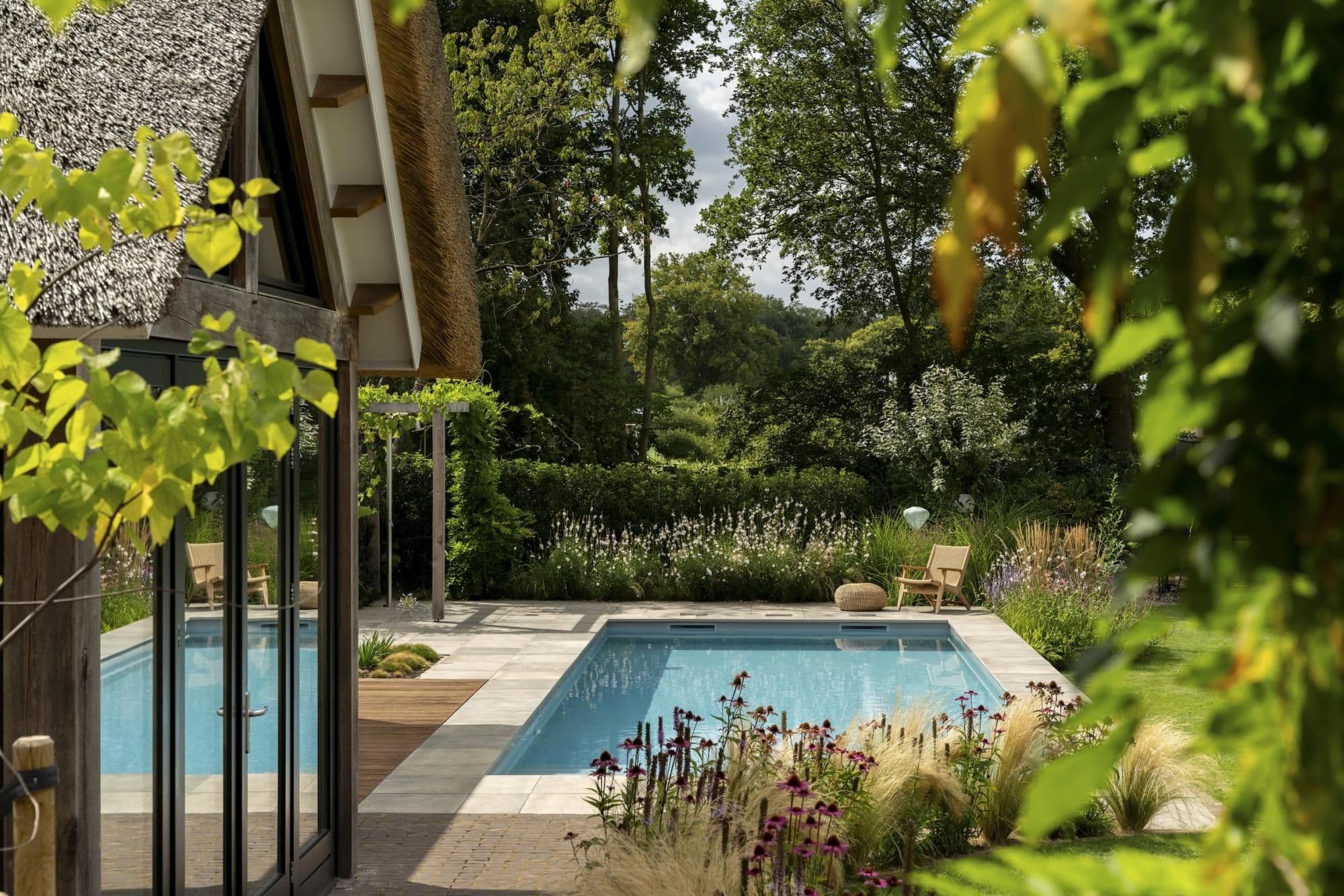 Rietgedekt huis met glazen schuifdeuren naast een zwembad op een betegeld terras, omgeven door bloeiende planten en bomen.