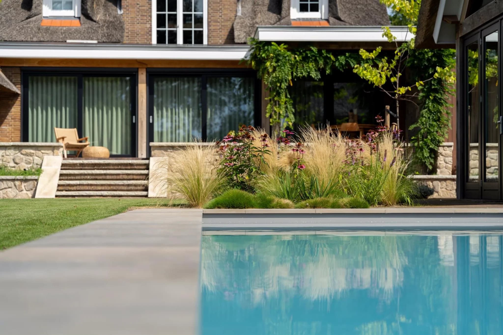 Zwembad op de voorgrond met reflectie, daarachter siergrassen en bloemen en een terras met trap voor een huis met rieten dak en schuifdeuren