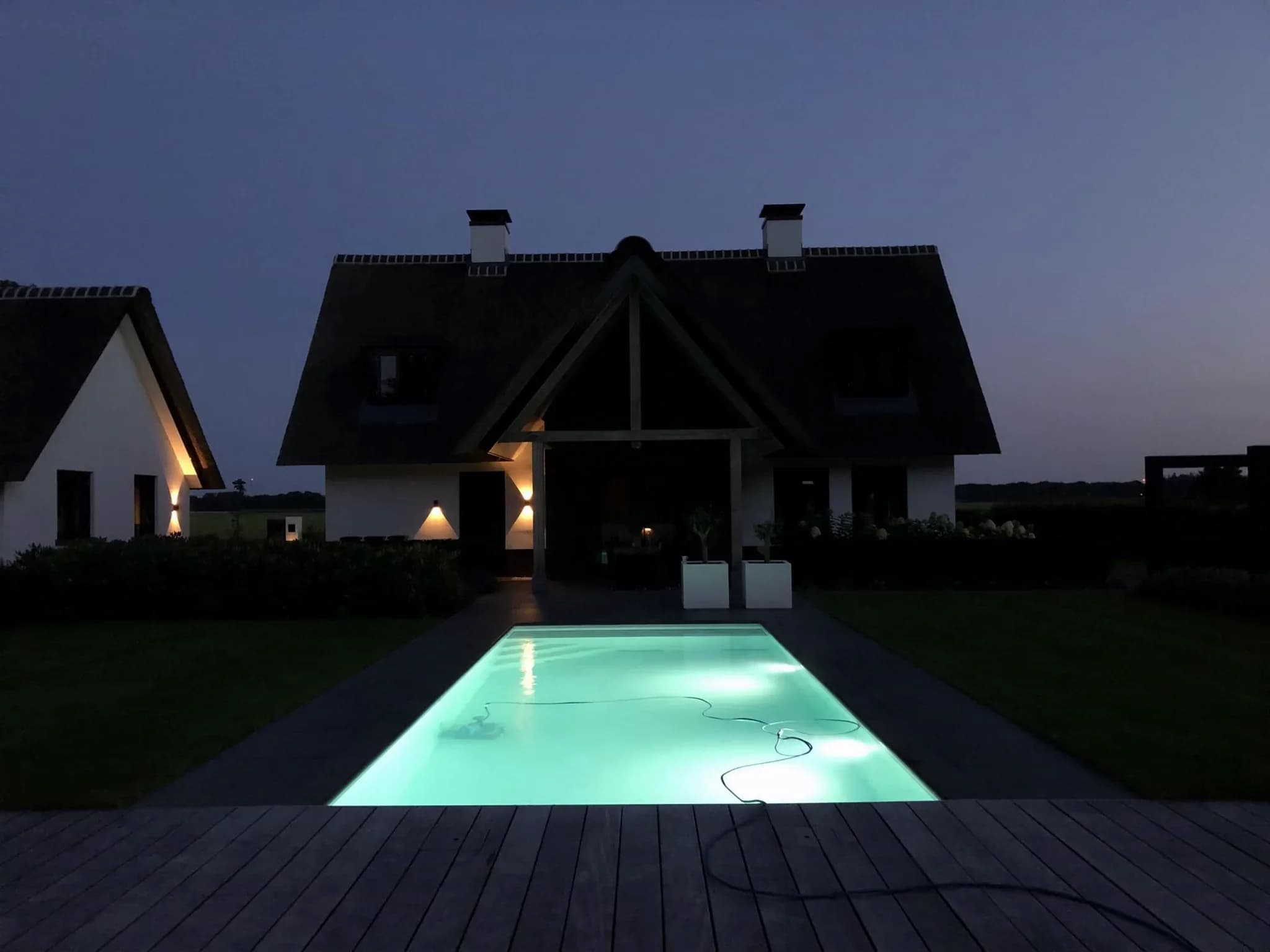 Voorzijde van een huis bij schemering met een verlicht zwembad op de voorgrond en wandlampen aan de gevel