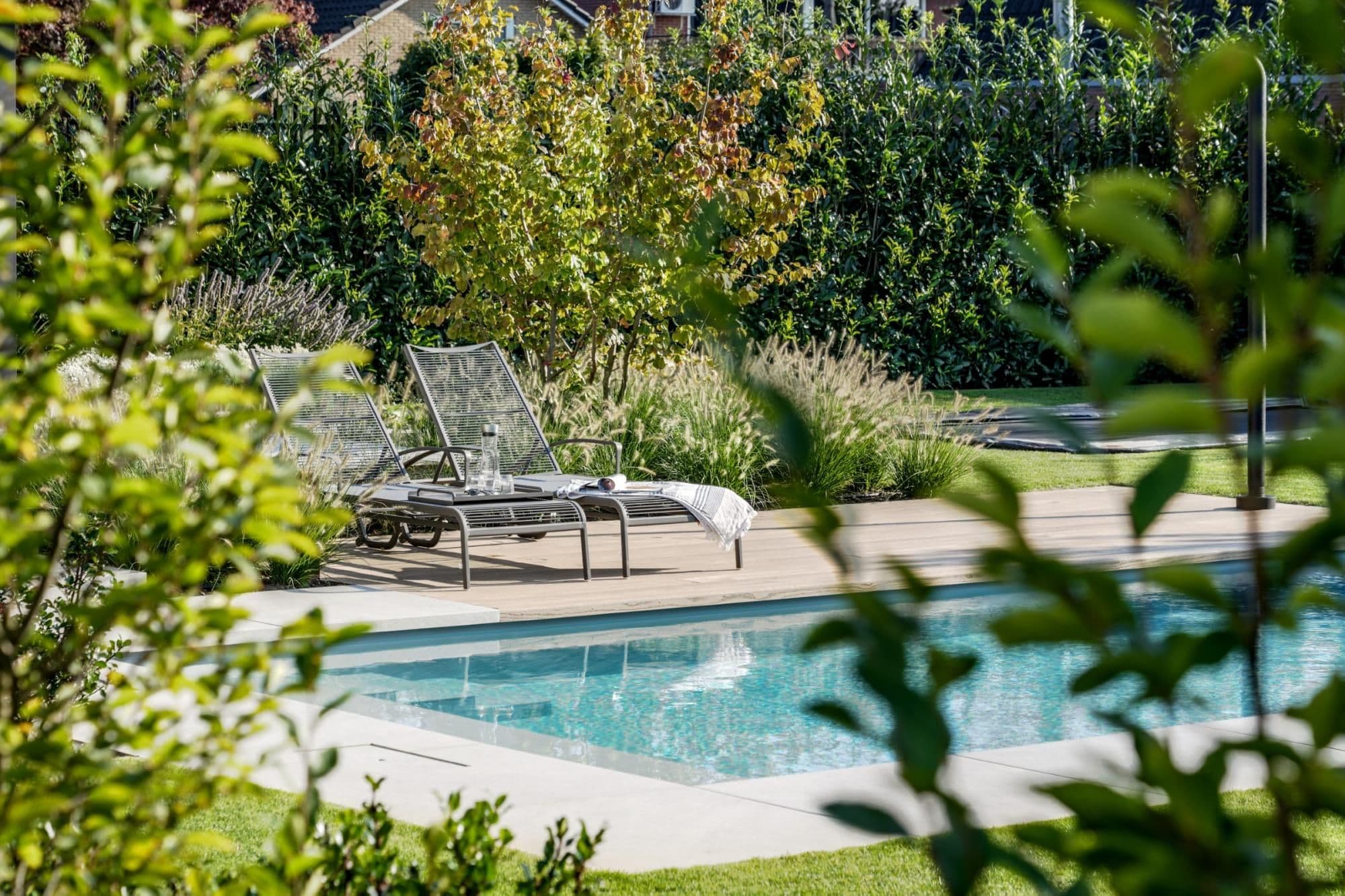 Twee metalen ligstoelen op een houten terras naast een zwembad, met een karaf en een handdoek, omgeven door gras, siergrassen en struiken