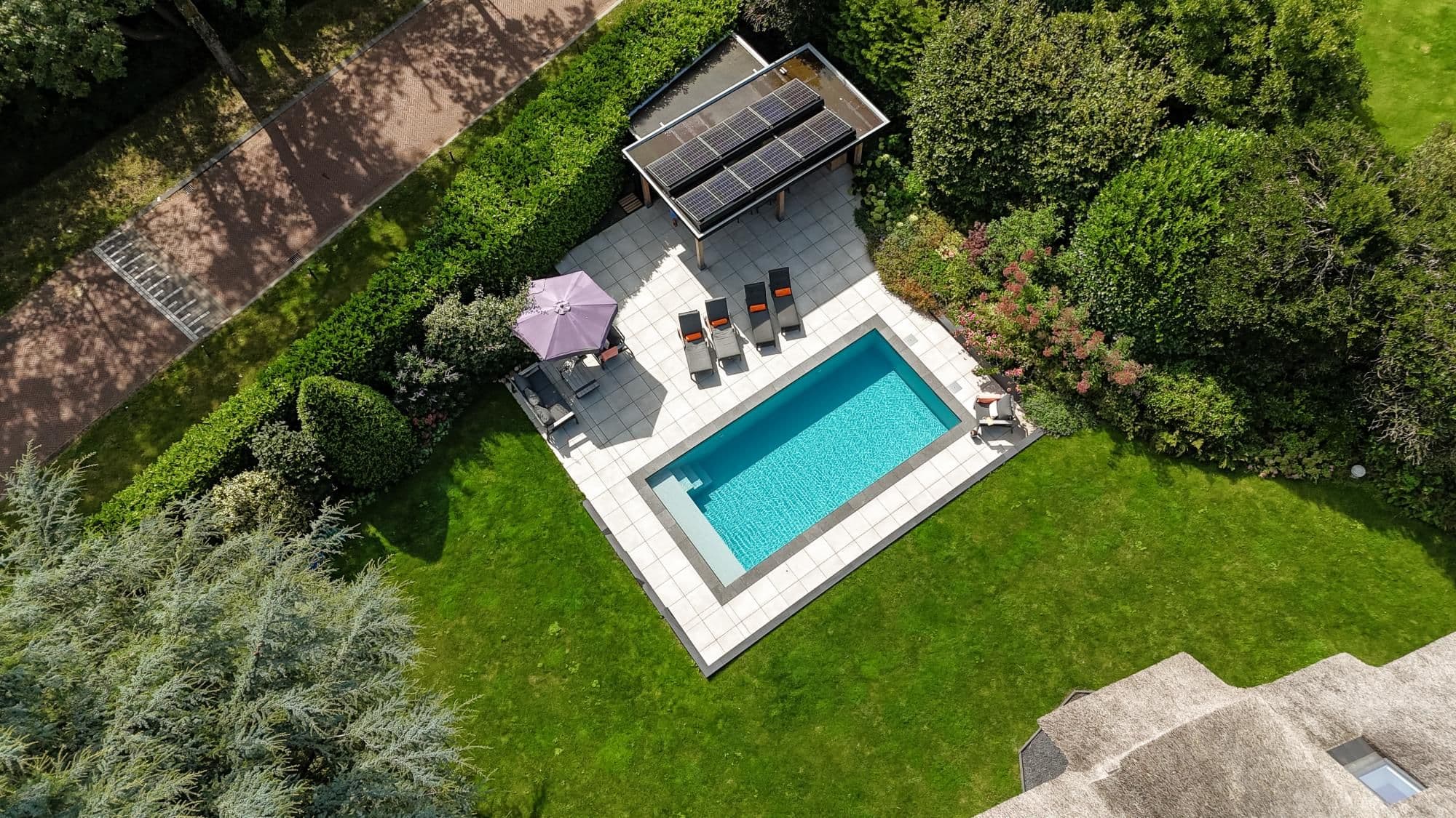 Luchtfoto van een achtertuin met een rechthoekig blauw zwembad, lichtkleurige terrastegels, vier ligstoelen met oranje kussens, een paarse parasol, een overkapping met zonnepanelen en omringend groen en gazon.