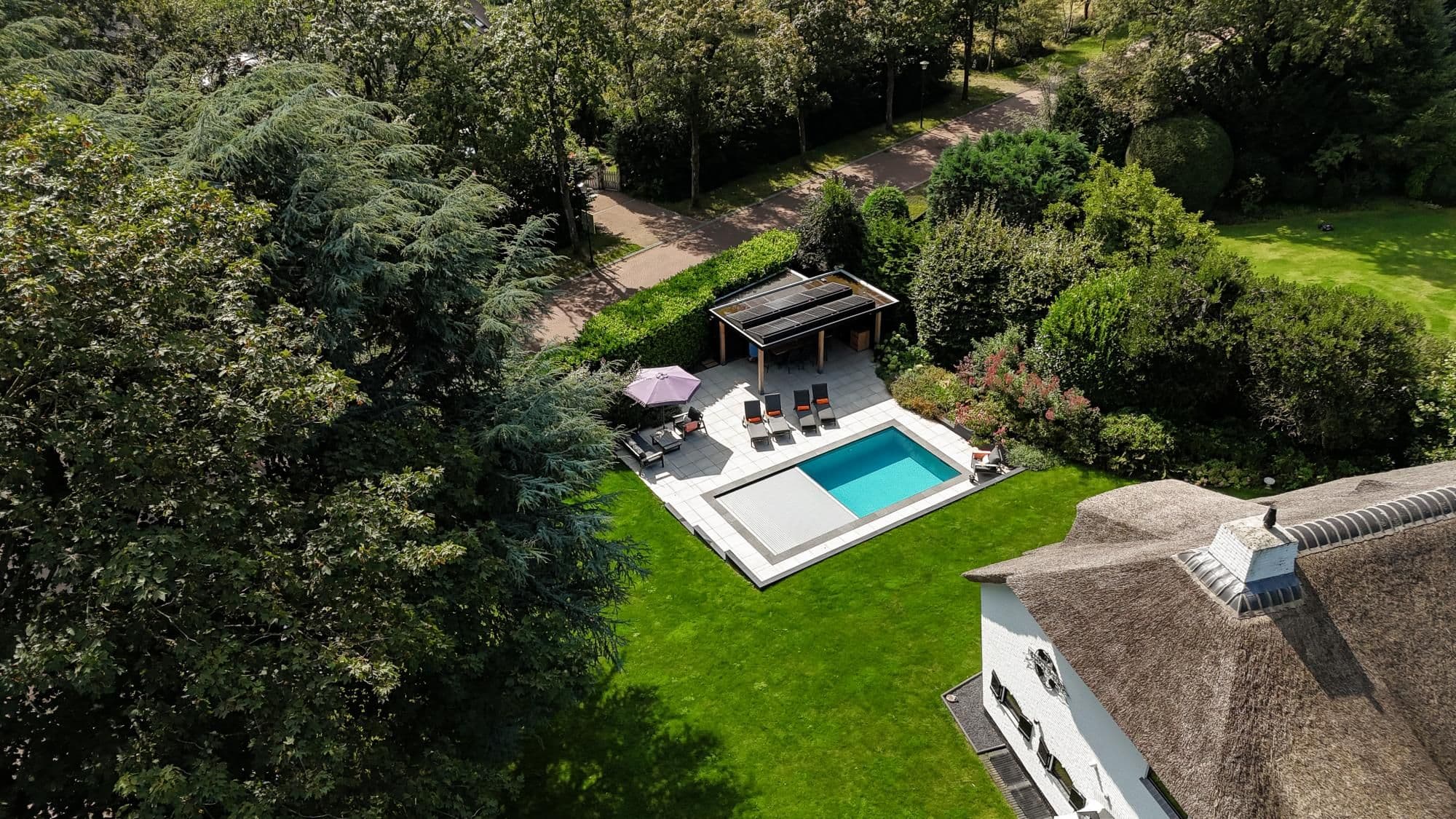 Luchtfoto van een besloten tuin met rechthoekig zwembad, terras met ligstoelen en parasol, overkapping met zonnepanelen en rieten dak zichtbaar