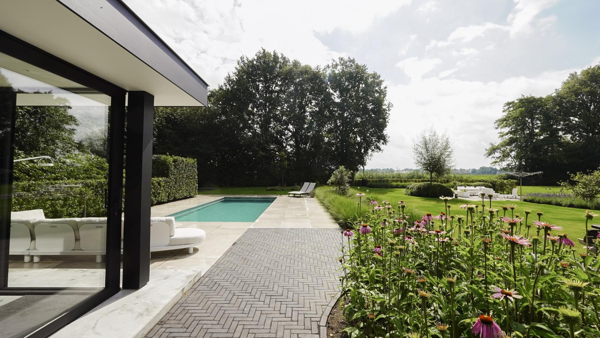 Terras en tuin van een modern huis met glazen schuifpui, ligstoelen, rechthoekig zwembad en paars-roze bloemen op de voorgrond