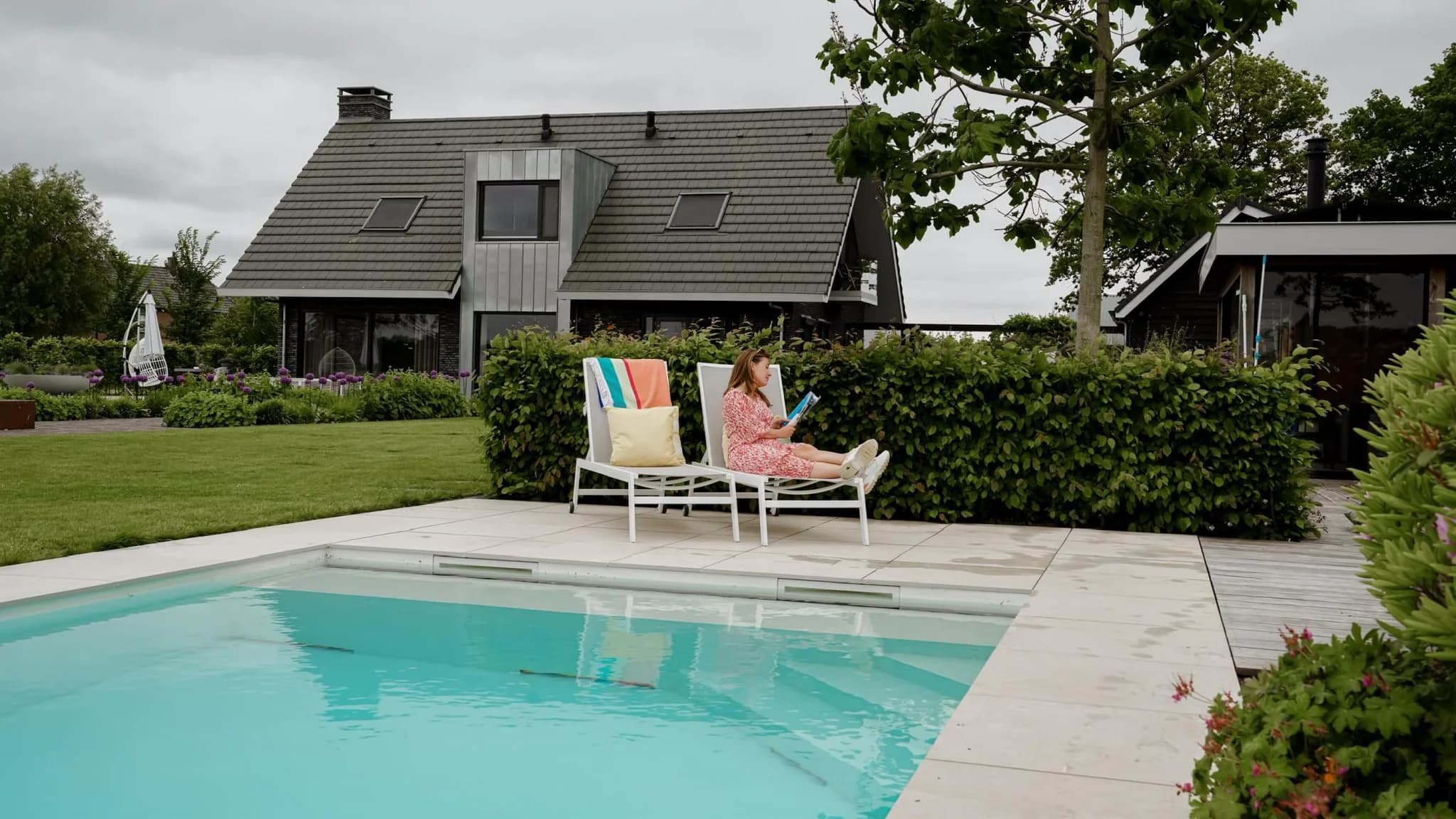 Persoon zit op een ligstoel naast een zwembad met een modern huis en groenblijvende hagen op de achtergrond