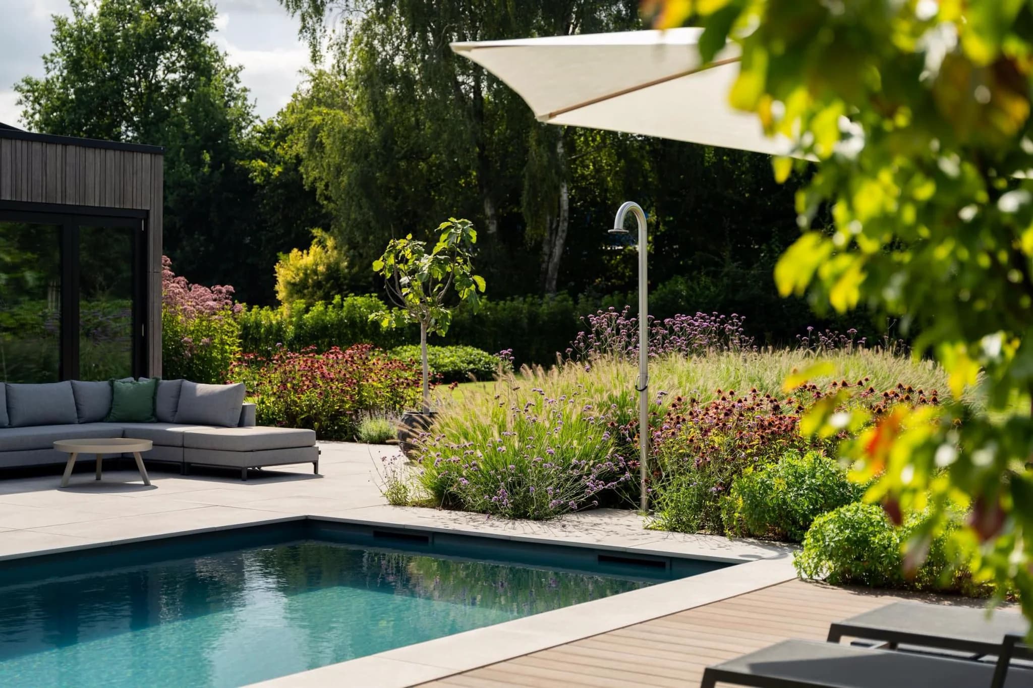 Zwembad en terras met grijze loungebank, parasol en buitendouche bij bloeiende borders