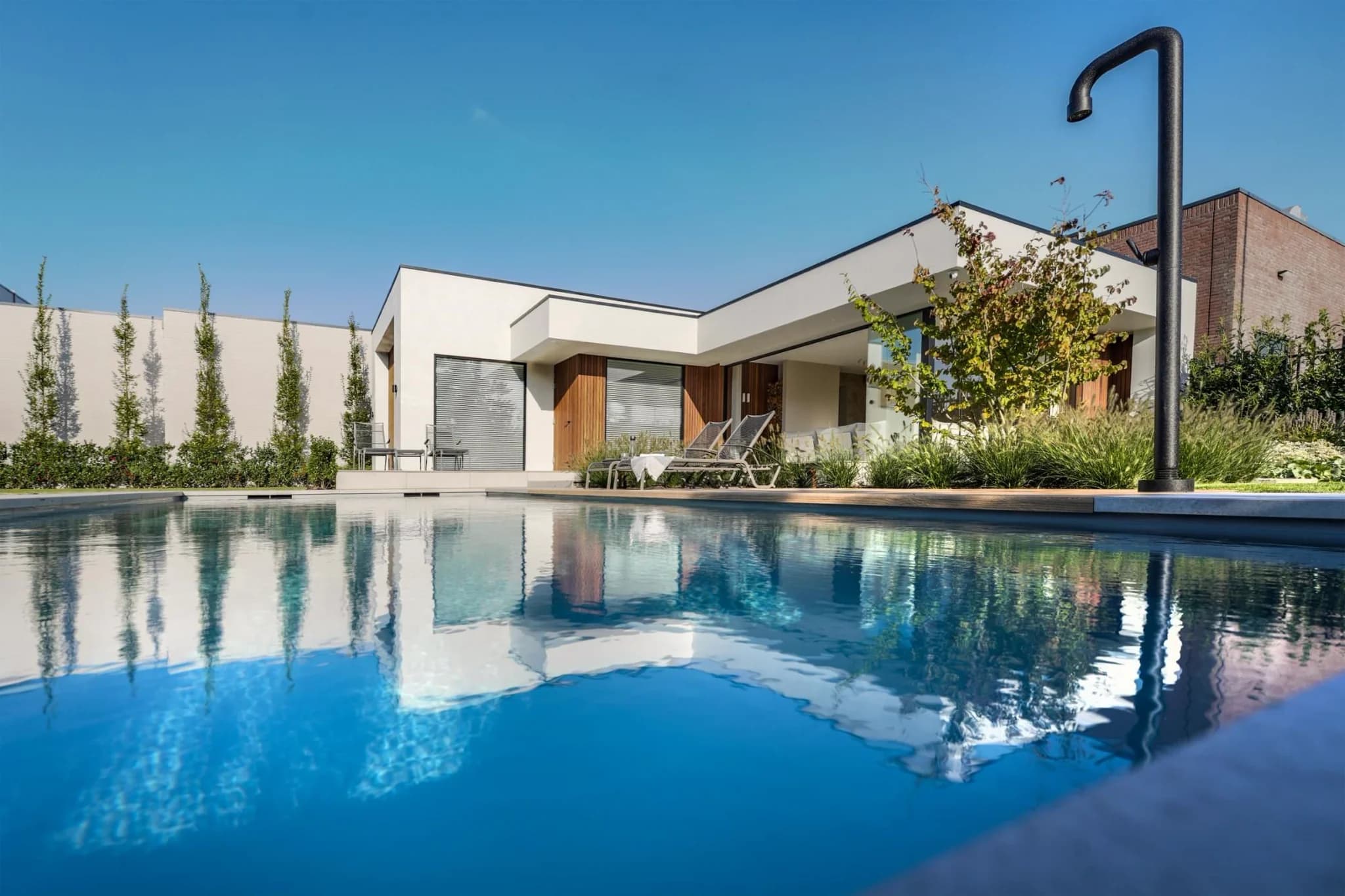 Moderne witte woning met houten accenten en ligstoelen naast een zwembad, met een zwarte buitendouche en reflectie in het water.