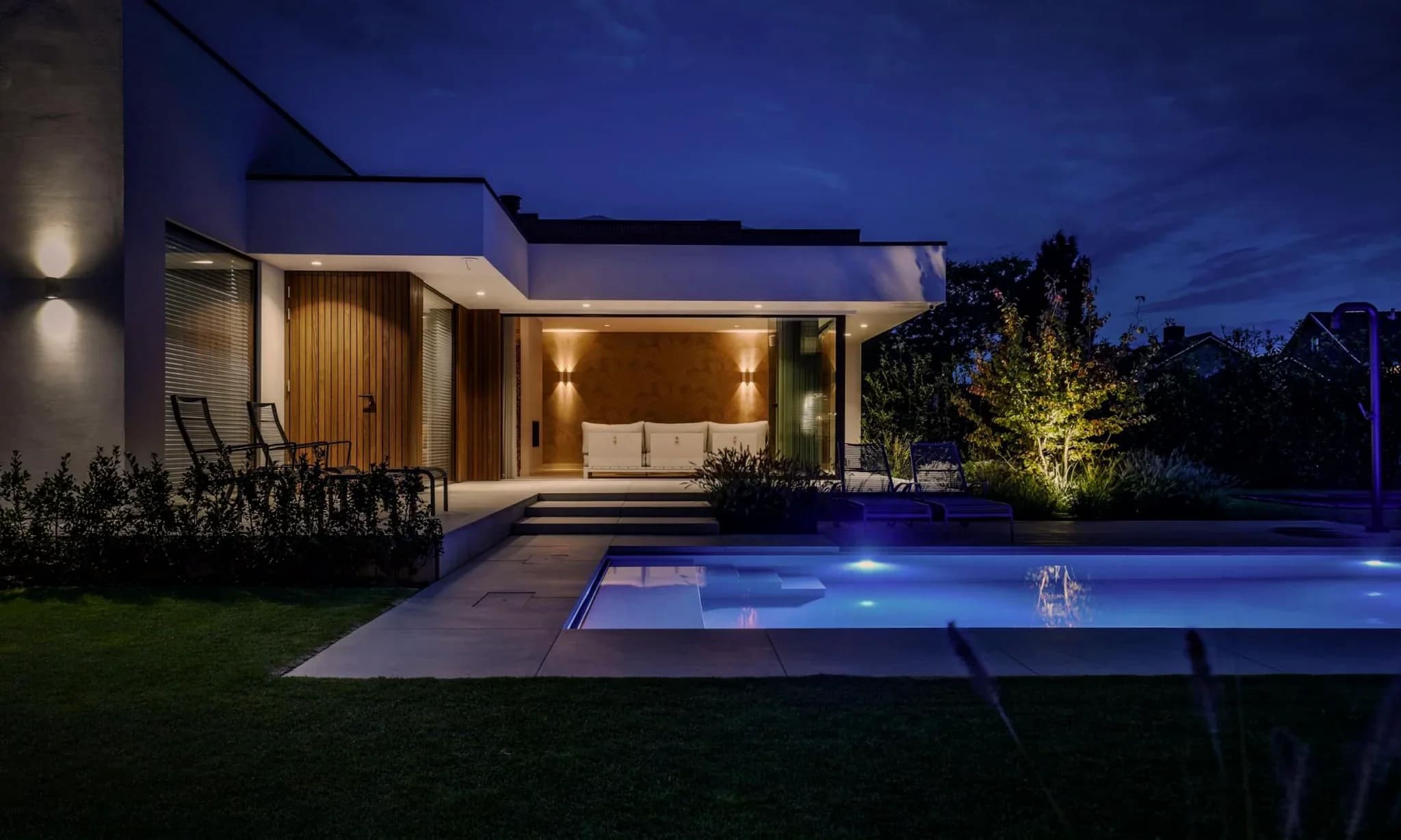 Voorgevel van een moderne woning met overdekte patio, houten accenten, buitenmeubilair en een verlicht zwembad in de avond