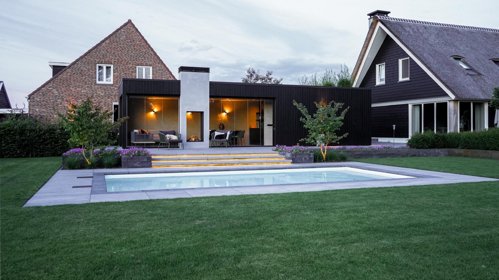 Voorgevel van moderne woning met zwembad, verhoogd terras met zitmeubilair en buitenhaard, verlichte treden en een hond op het terras