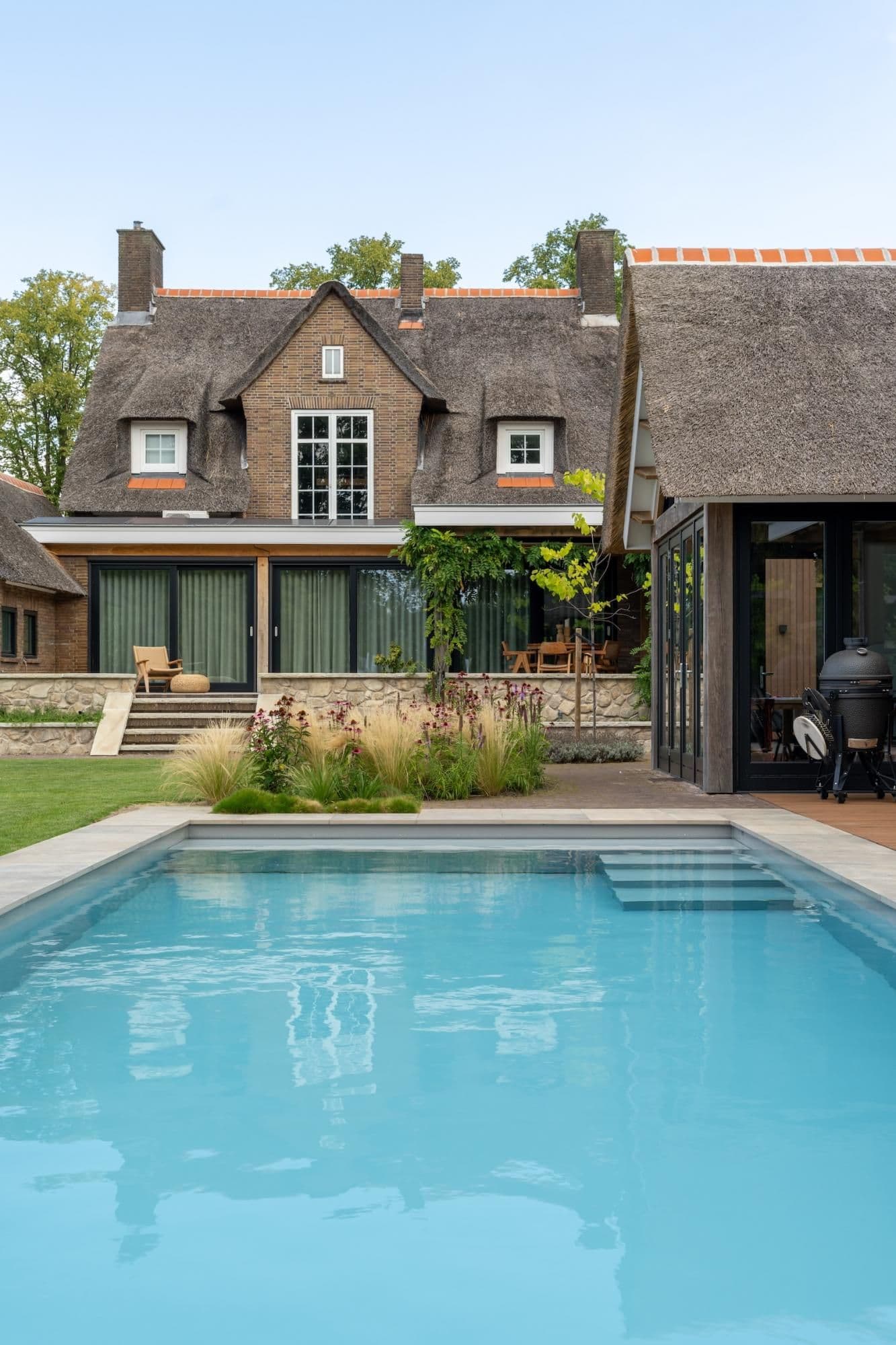 Voorgevel van een rietgedekte woning met grote glazen schuifdeuren, terras, beplanting en een blauw zwembad op de voorgrond.