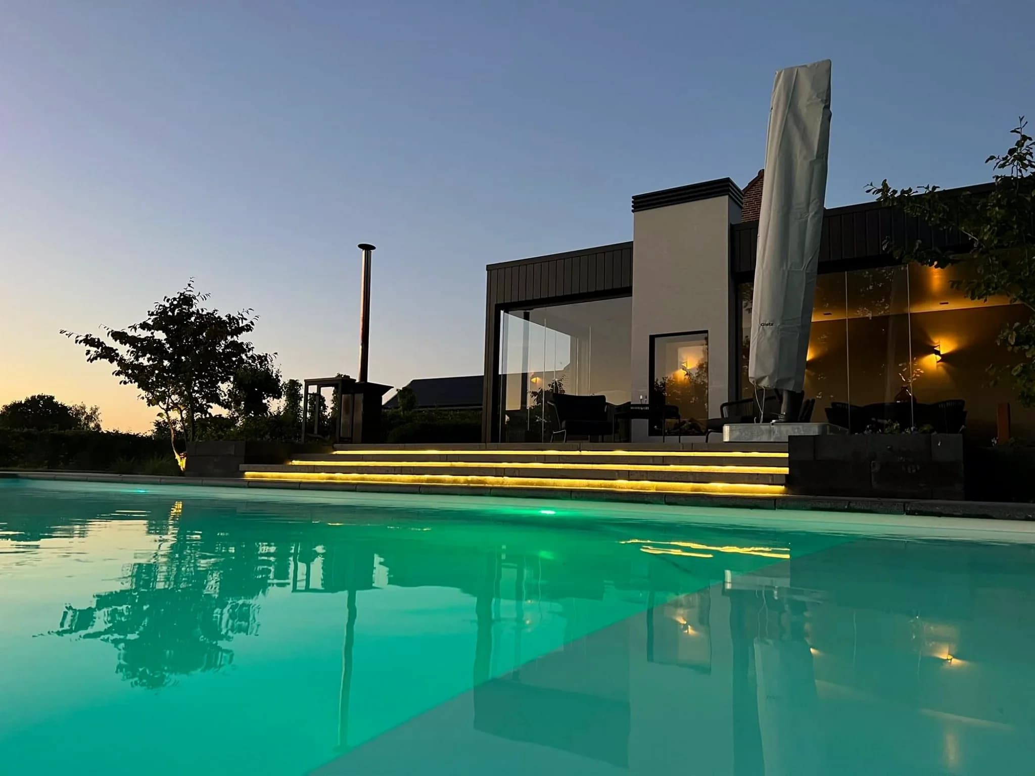 Modern terras met glazen gevel, overdekt zitgedeelte, verlichte trap en zwembad op de voorgrond bij schemering
