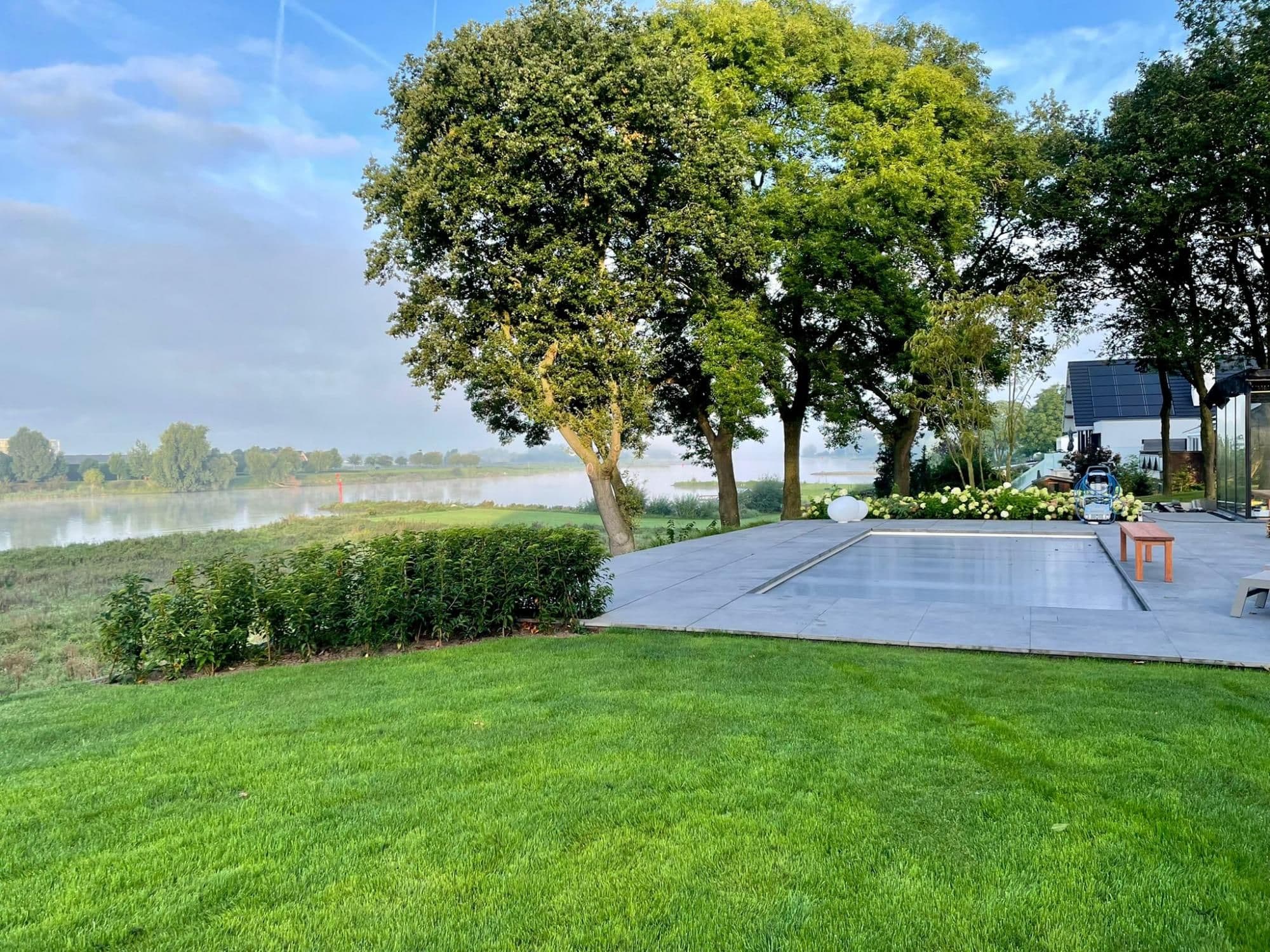 Groen gazon op de voorgrond, rechts een betegeld terras met afgedekt zwembad, achteraan bomen en een rivier.