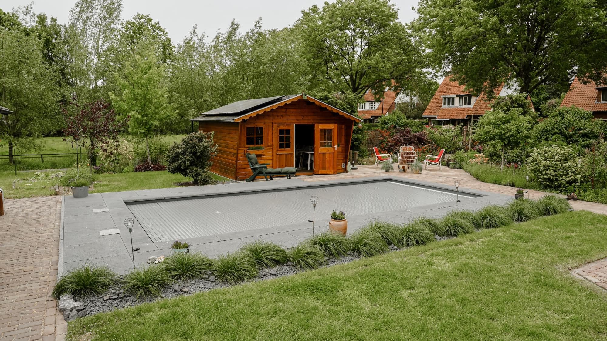 Tuin met een overdekt zwembad op een betegeld terras, een houten tuinhuis met zonnepanelen op het dak en tuinmeubels tussen groenblijvende beplanting.