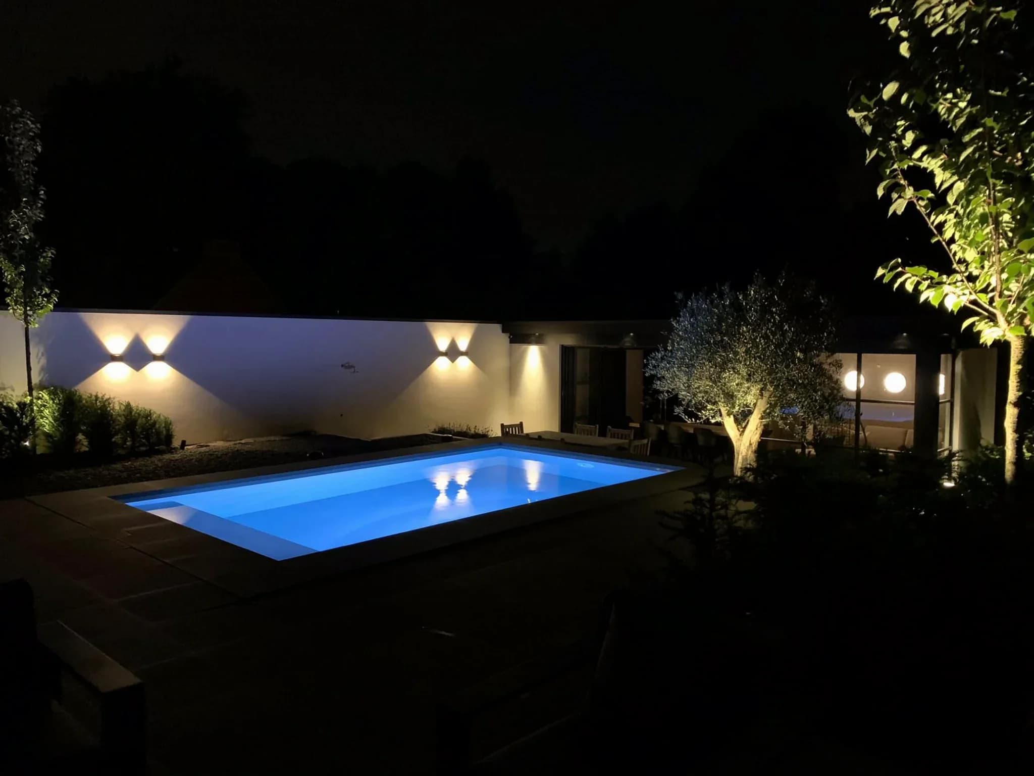 Buitenzwembad met blauwe verlichting, verlichte muur en tuinverlichting in een tuin bij nacht