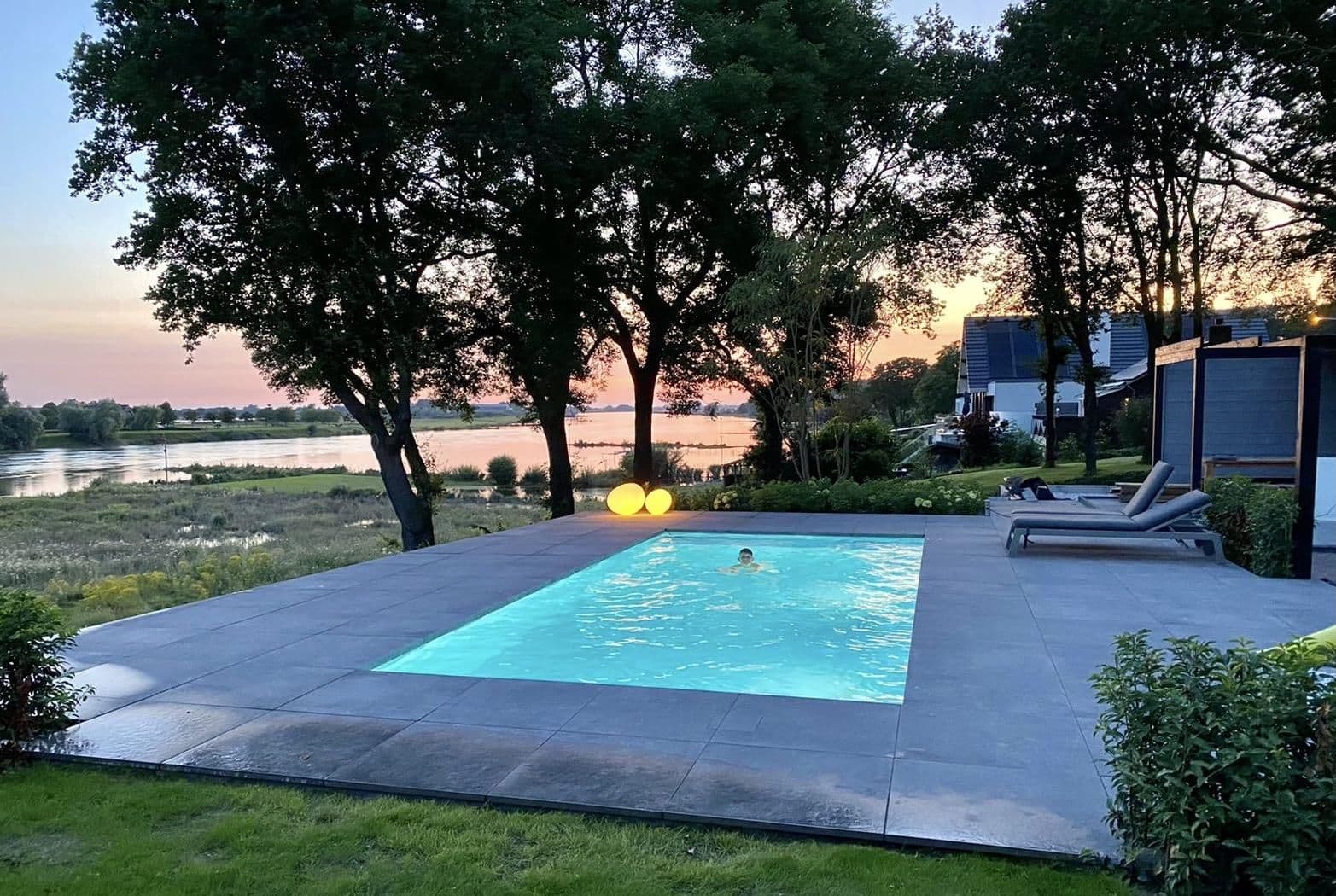 Rechthoekig verlicht zwembad met een persoon zwemmend, betegeld terras, ligstoelen en bomen met uitzicht op een rivier bij zonsondergang