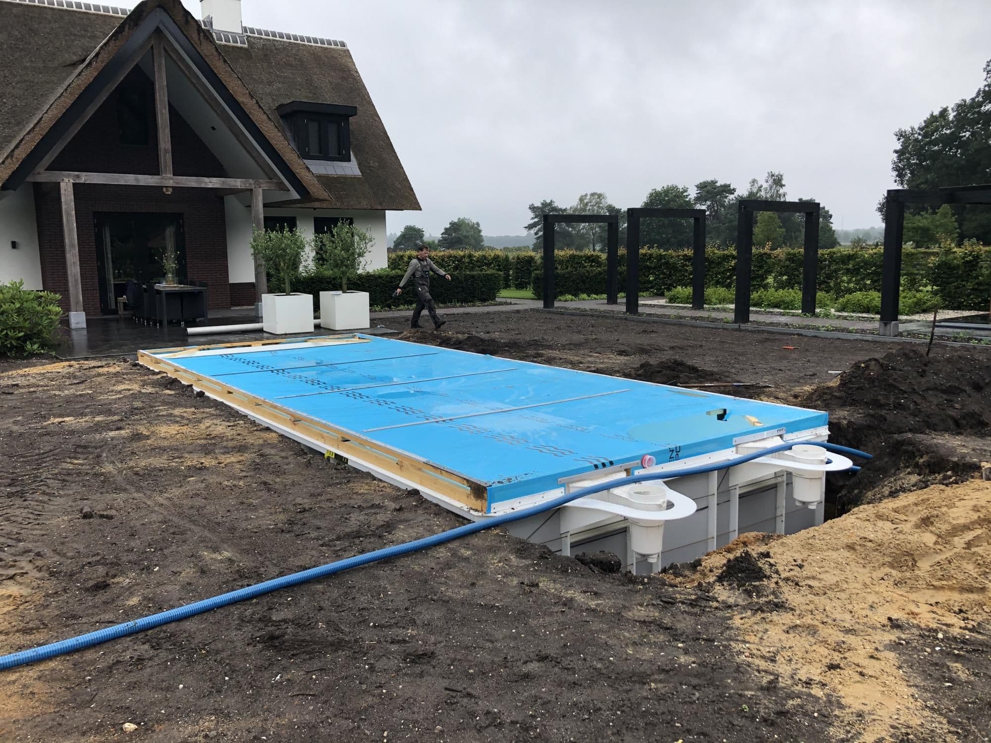 Woning met rietdak, blauwe platen en kunststof zwembadonderdelen in de voortuin, persoon lopend en pergola-frames op de achtergrond