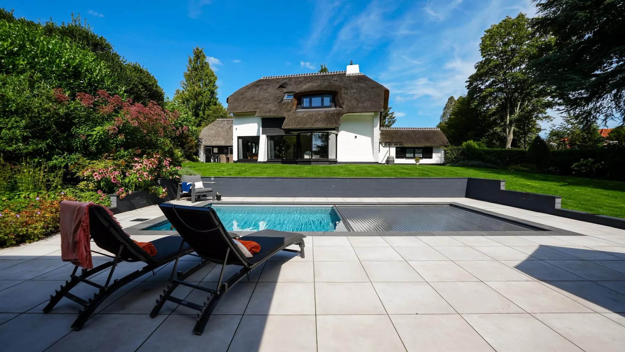Voorzijde van een witte woning met rieten dak, grasveld, planten en een zwembad met ligstoelen op een betegeld terras.