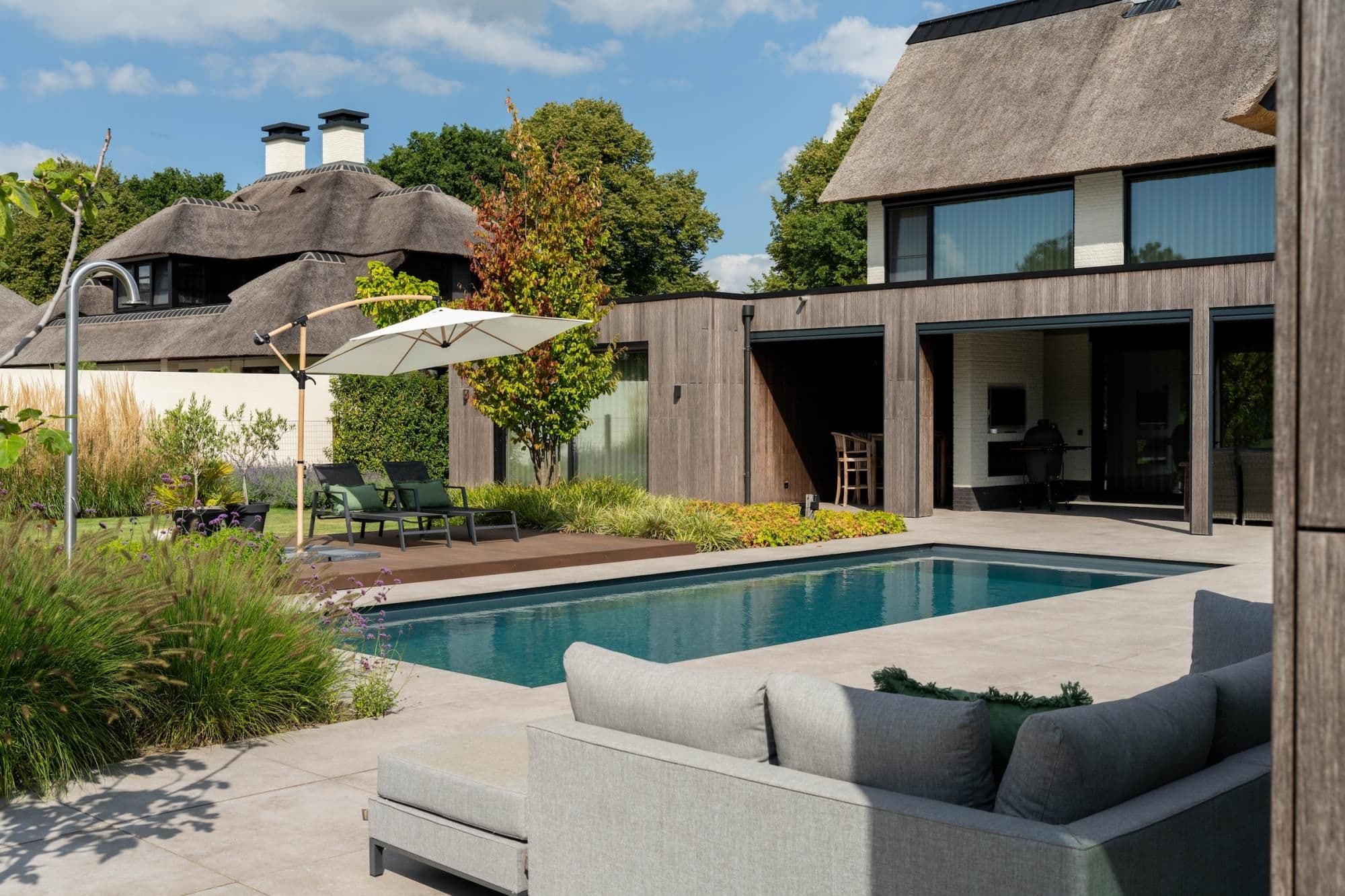 Voorzijde van een moderne woning met rieten dak, houten gevel, zwembad, ligstoelen en parasol