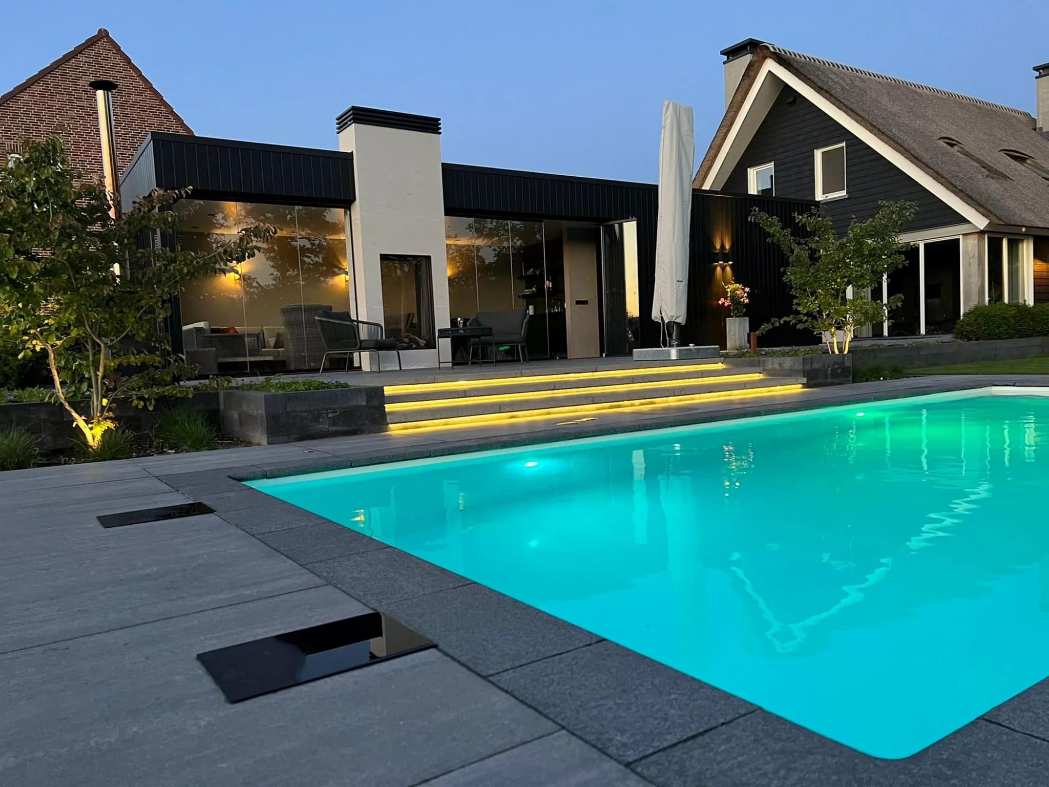 Woning bij schemering met helder verlicht zwembad, traptreden met gele verlichting en terras met glazen gevel en stoelen