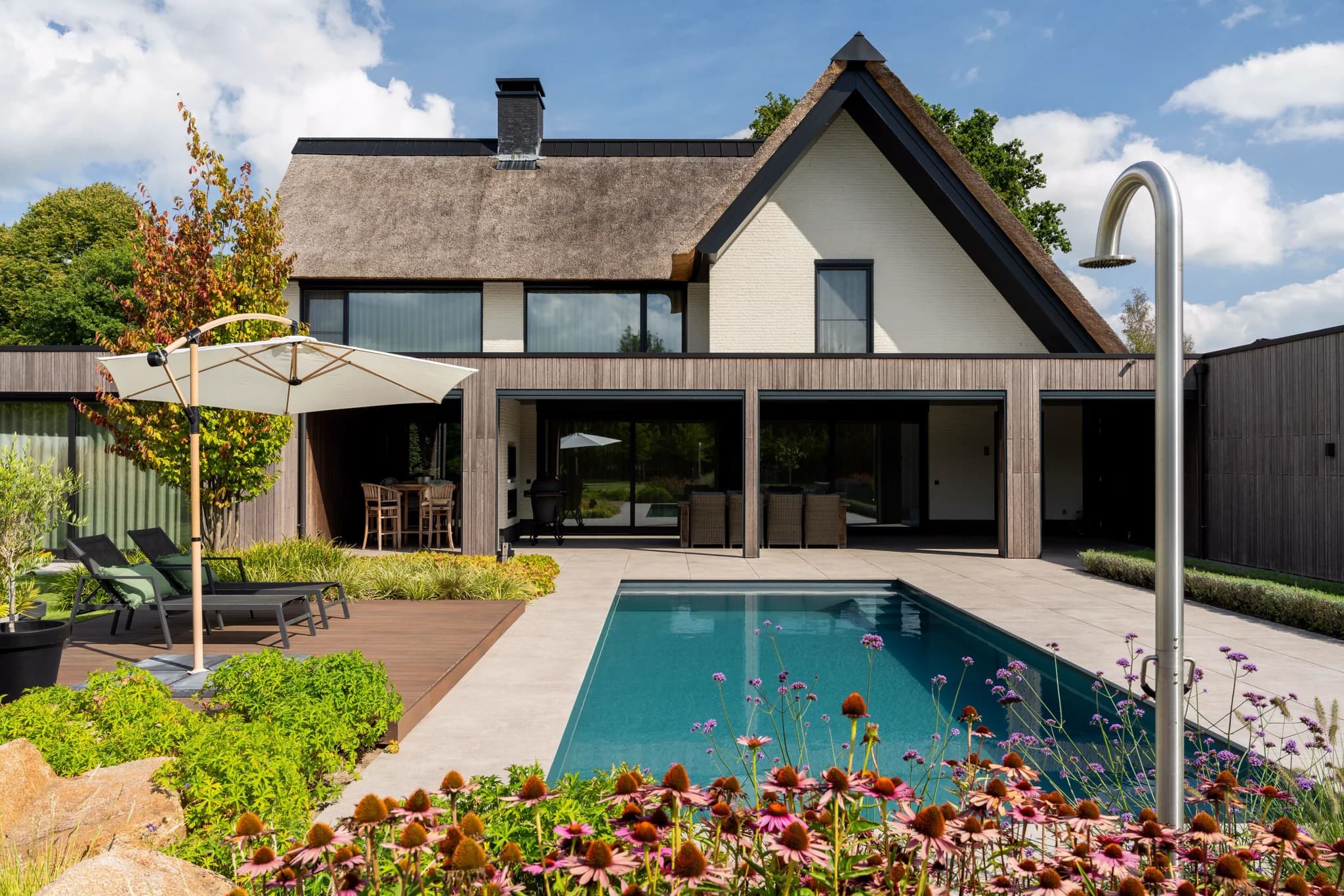 Voorzijde van huis met rieten dak, houten gevel, terras met parasol en ligstoelen, zwembad en roestvrijstalen buitendouche