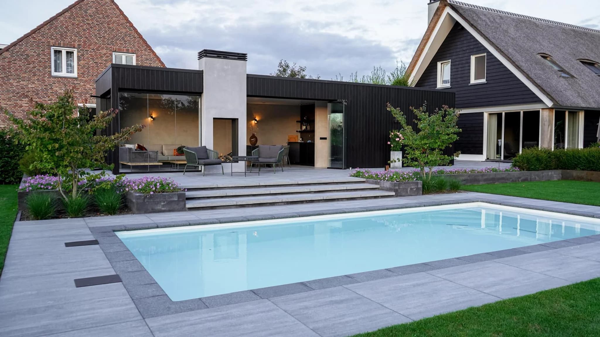 Woonhuis met zwarte uitbouw en overdekt terras met loungezetels, planten en een rechthoekig buitenzwembad op de voorgrond.