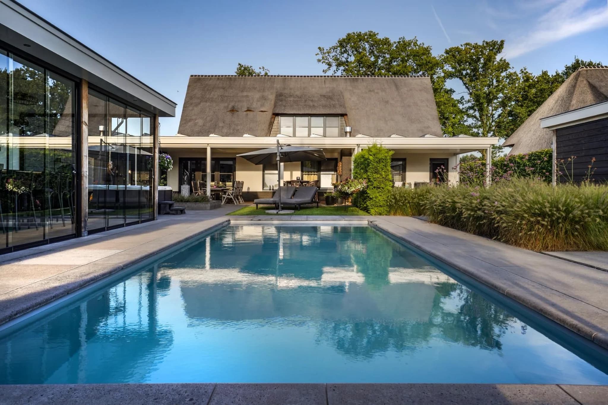Voorzijde van een woonhuis met overdekt terras, ligbedden, parasol en een rechthoekig zwembad op de voorgrond