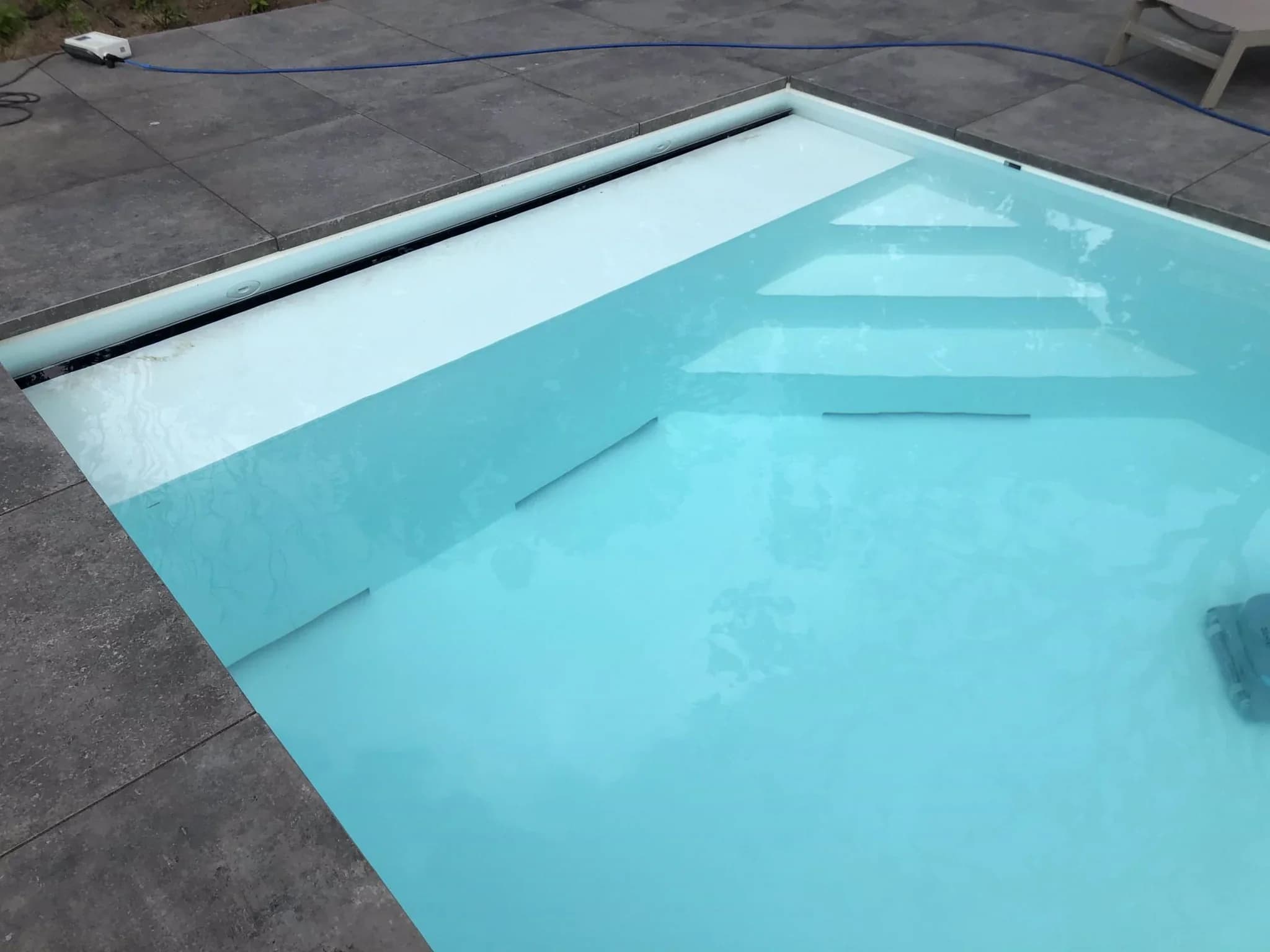 Bovenzijde van een zwembad met lichtblauw water, ingebouwde brede treden en grijze terrasstenen; wit apparaat en blauwe kabel op het terras.