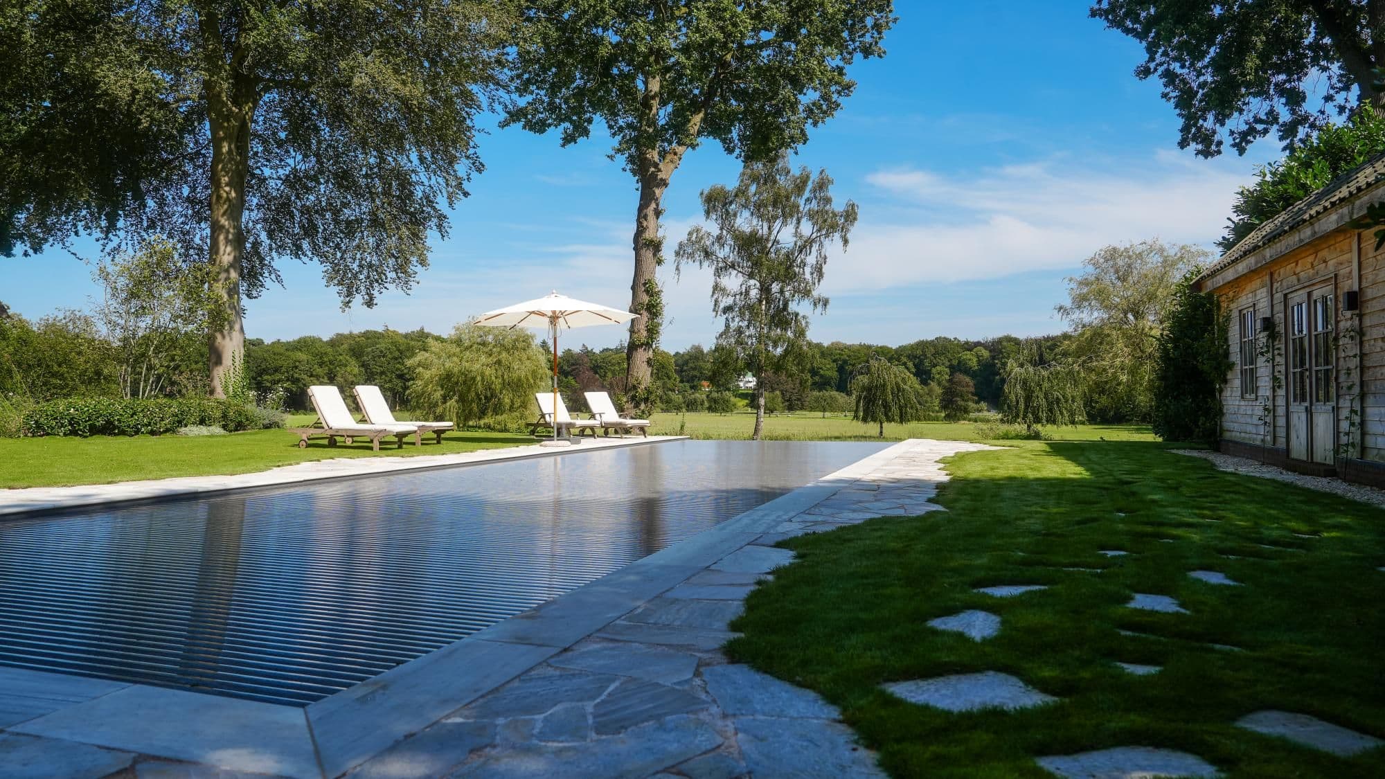 Zwembad met bedekking op de voorgrond, vier ligstoelen en een witte parasol op gras, bomen op de achtergrond en een houten bijgebouw rechts