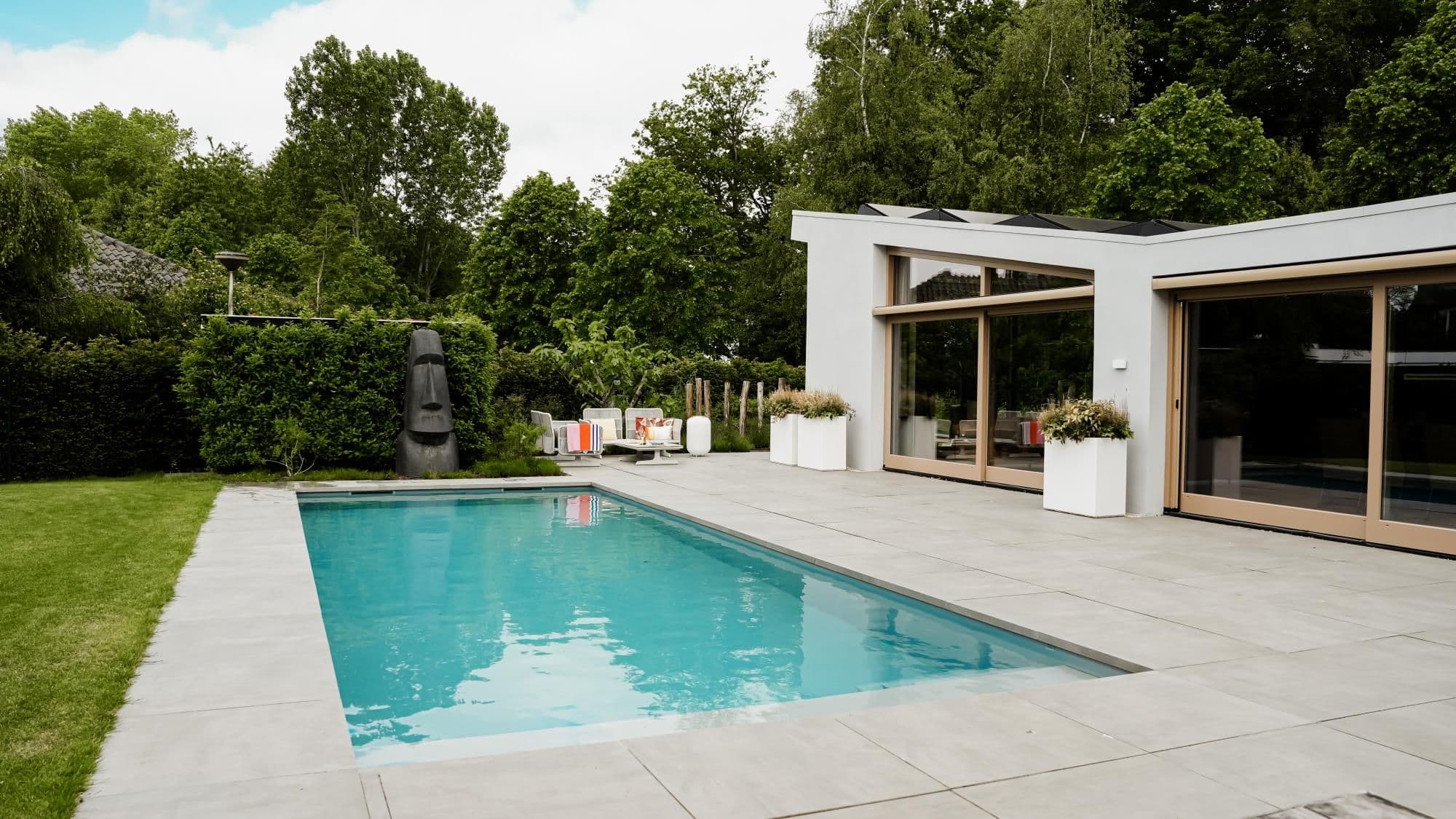 Rechthoekig buitenzwembad naast modern wit huis met betegeld terras, plantenbakken, lounge stoelen en een grote stenen sculptuur, omgeven door groen.