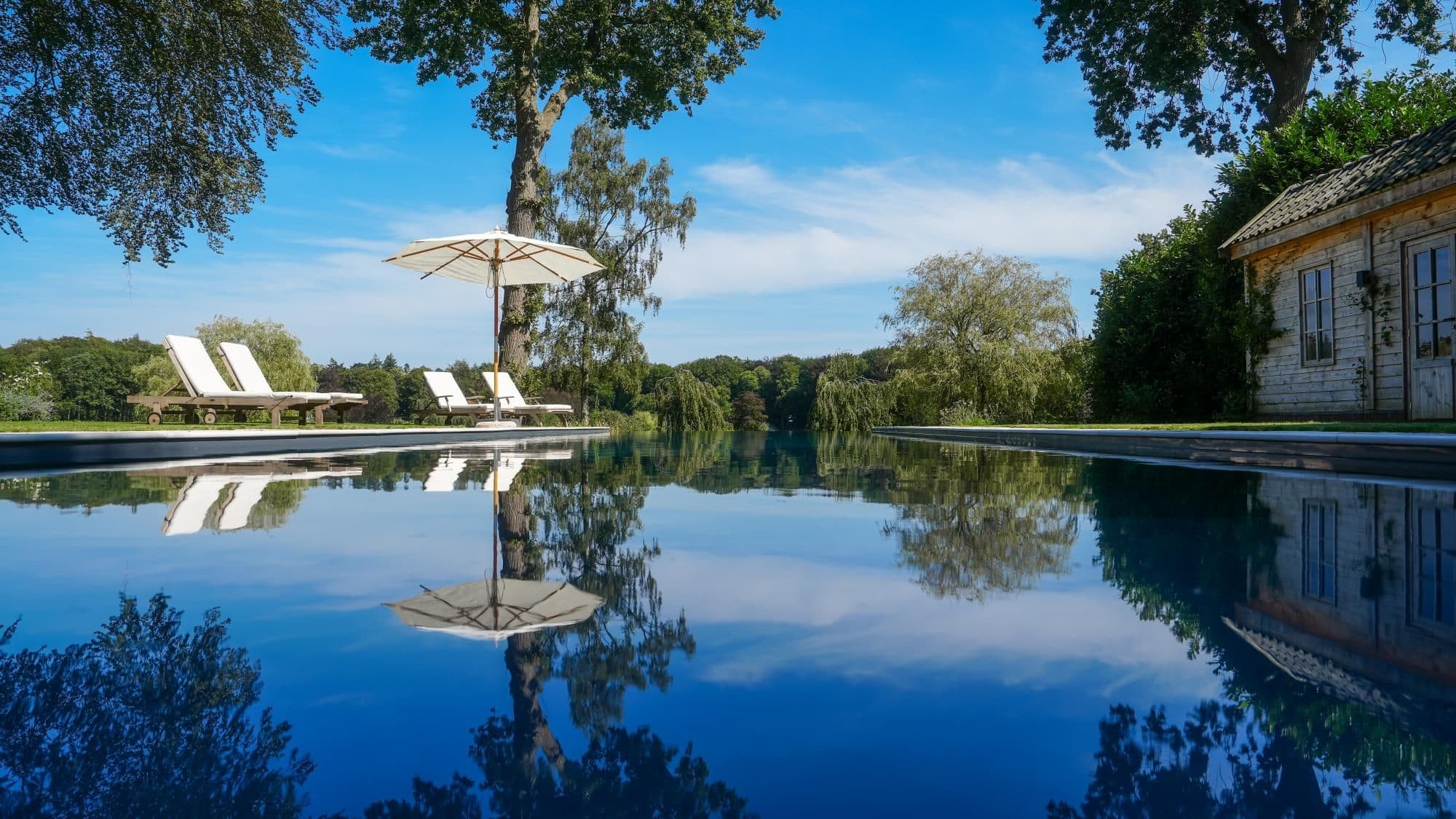 Zwembad met spiegelend water waarop een parasol, twee ligstoelen en bomen worden weerspiegeld; rechts een houten tuinhuis en op de achtergrond groen landschap onder een blauwe lucht.