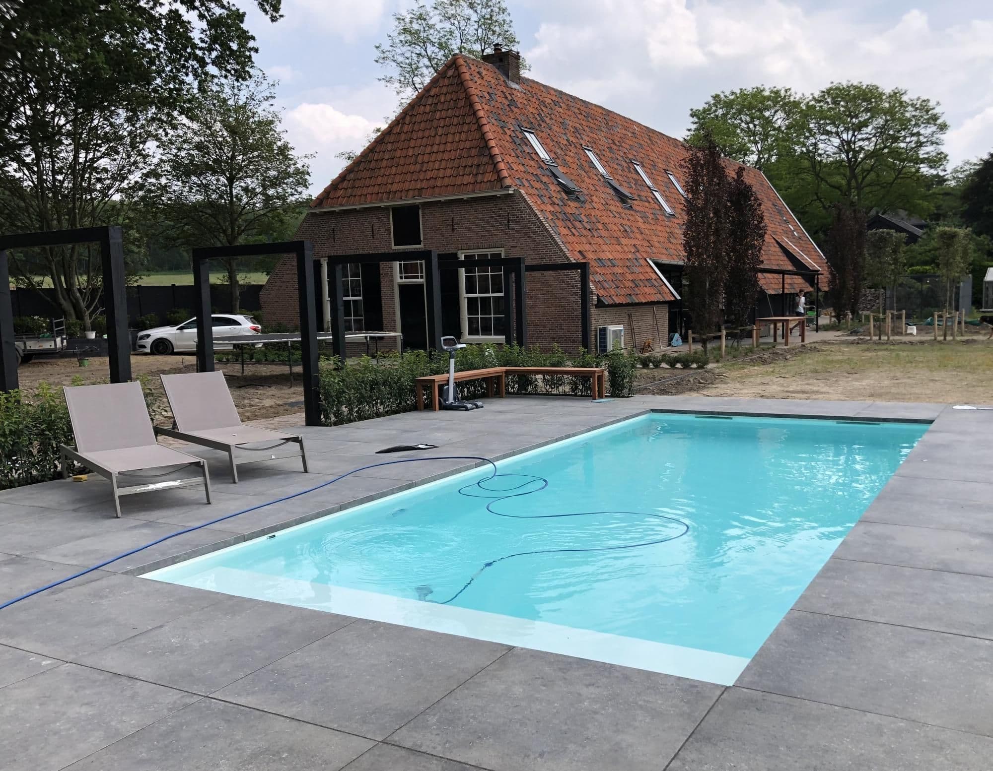 Zwembad met blauwe water, schoonmaakslang in het water, ligstoelen op betegeld terras en bakstenen woonhuis met rood pannendak op de achtergrond