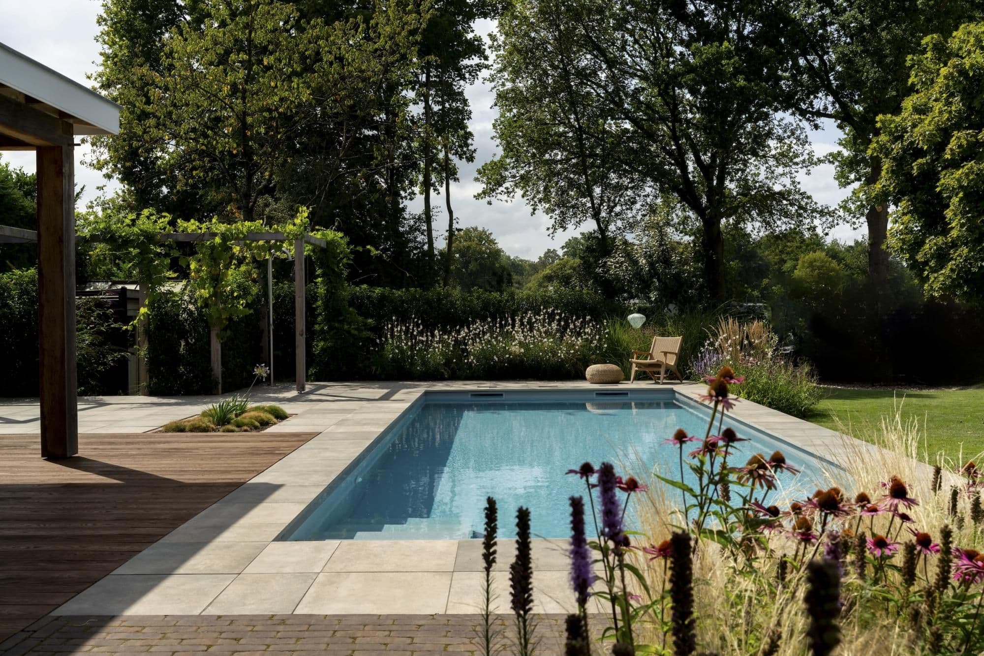 Rechthoekig zwembad met blauw water, betegeld terras en houten vlonder, links een pergola, rechts een rieten stoel en poef bij bloemen en siergrassen, bomen op de achtergrond