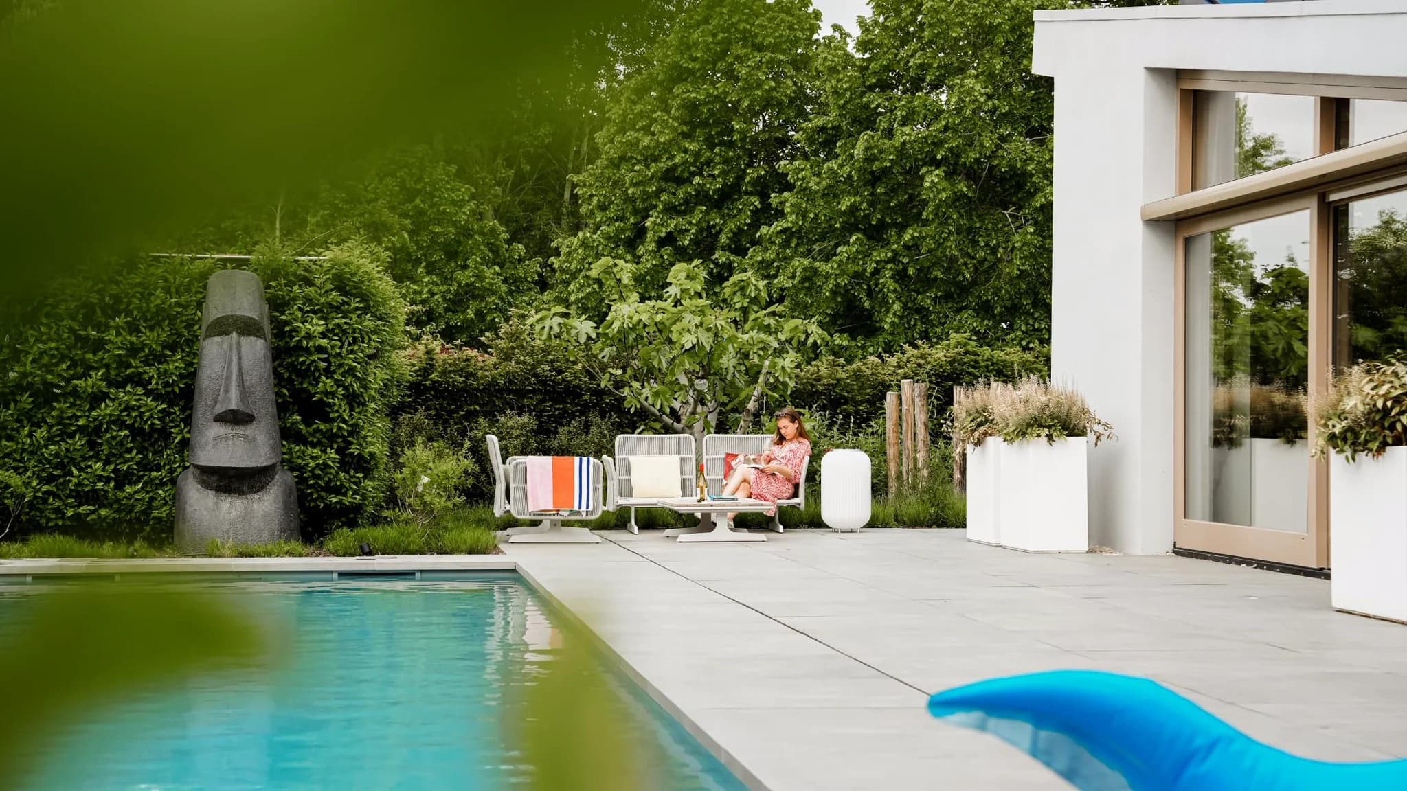 Terras bij zwembad met loungezitjes, een vrouw die op een stoel leest, een groot stenen beeld en veel groen naast een moderne woning