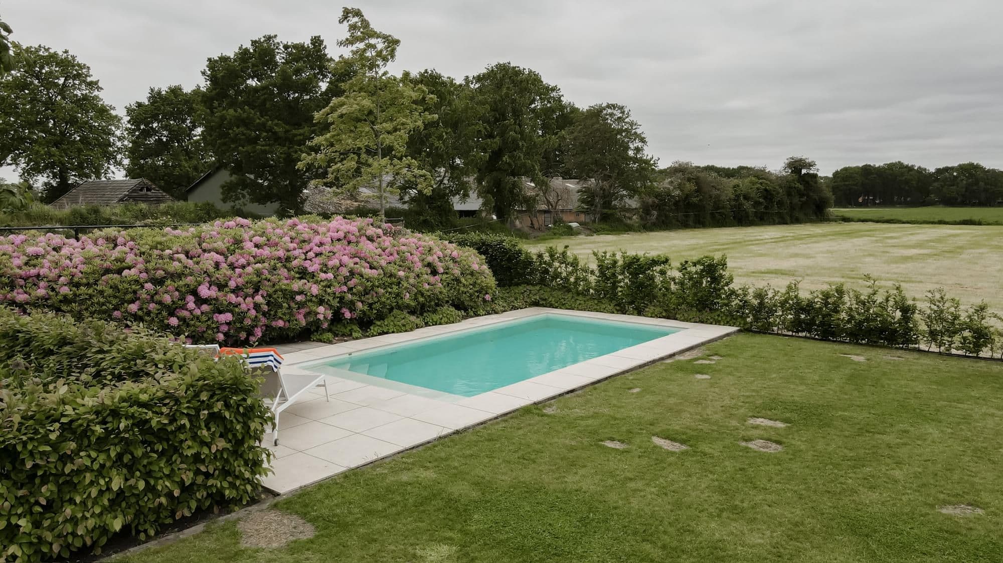 Rechthoekig zwembad met blauw water op betegeld terras, omgeven door hagen en roze rododendrons, met weiland en boerderijgebouwen op de achtergrond onder een bewolkte lucht.