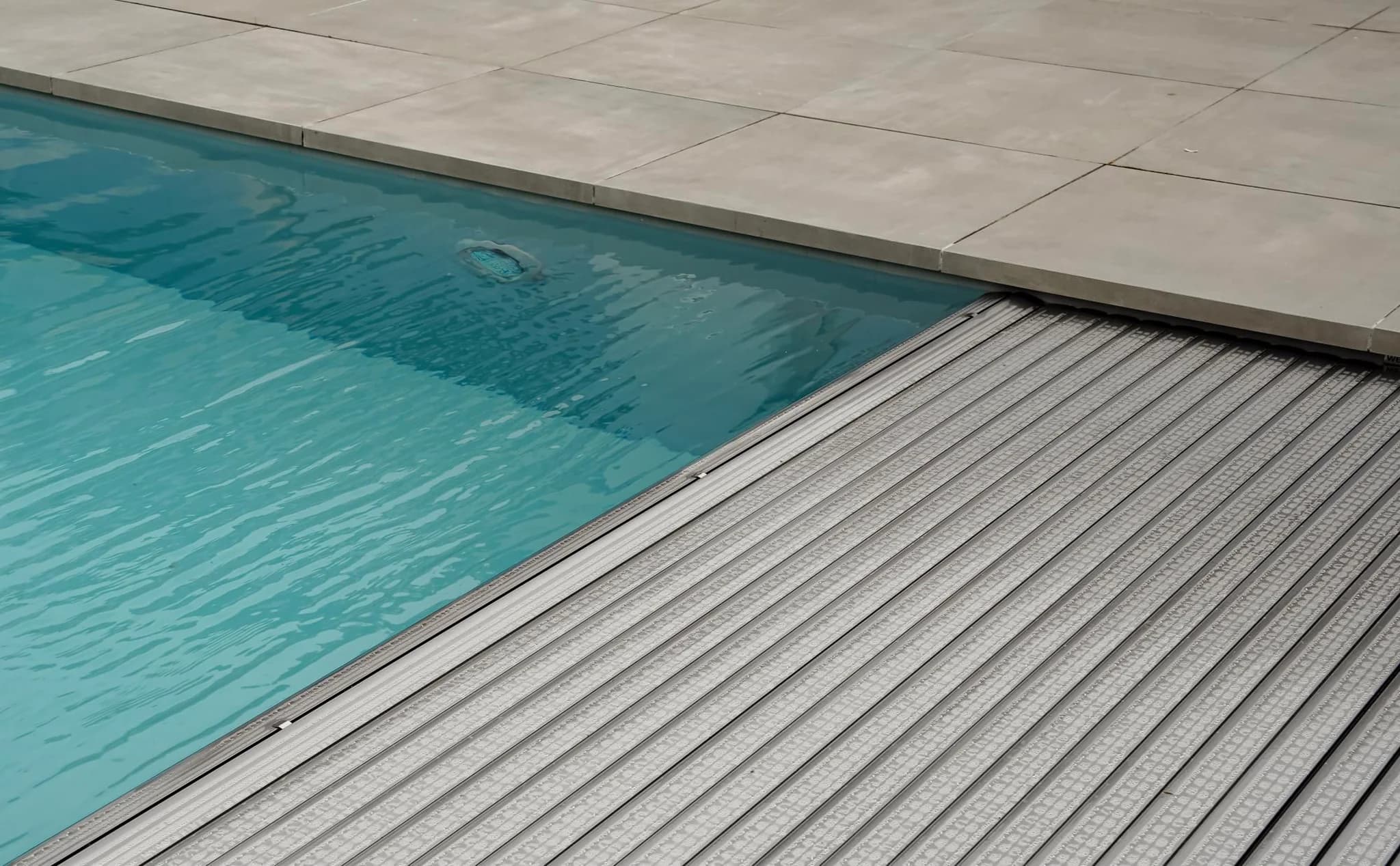 Zwembadrand met blauw water, lichtgrijze tegels en een gedeeltelijk zichtbare geribde afdekking langs de waterlijn. Ronde inbouwunit onder water zichtbaar.