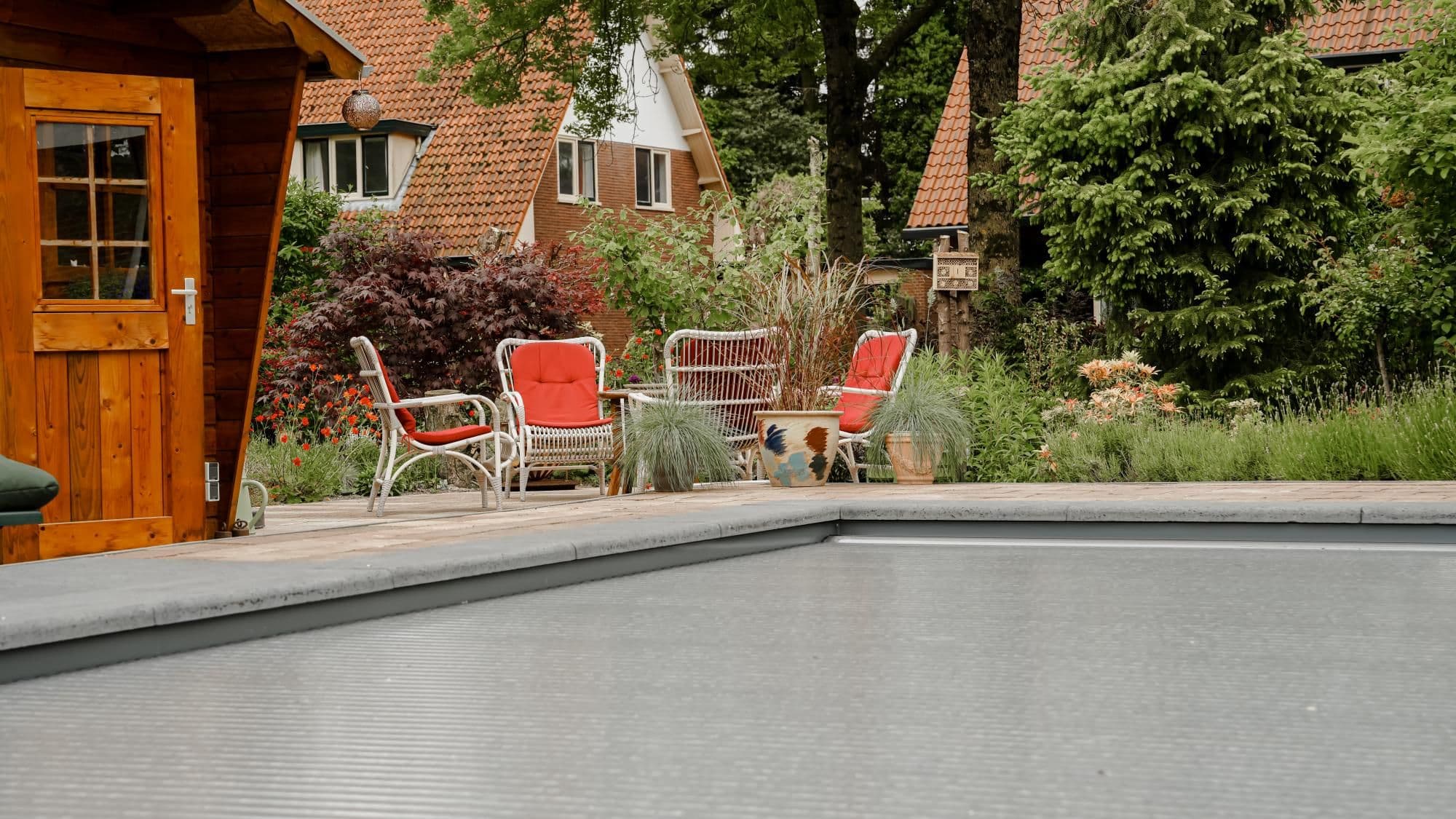 Tuin met houten tuinhuis, gevlochten stoelen met rode kussens, diverse planten en een gesloten zwembadafdekking op de voorgrond.
